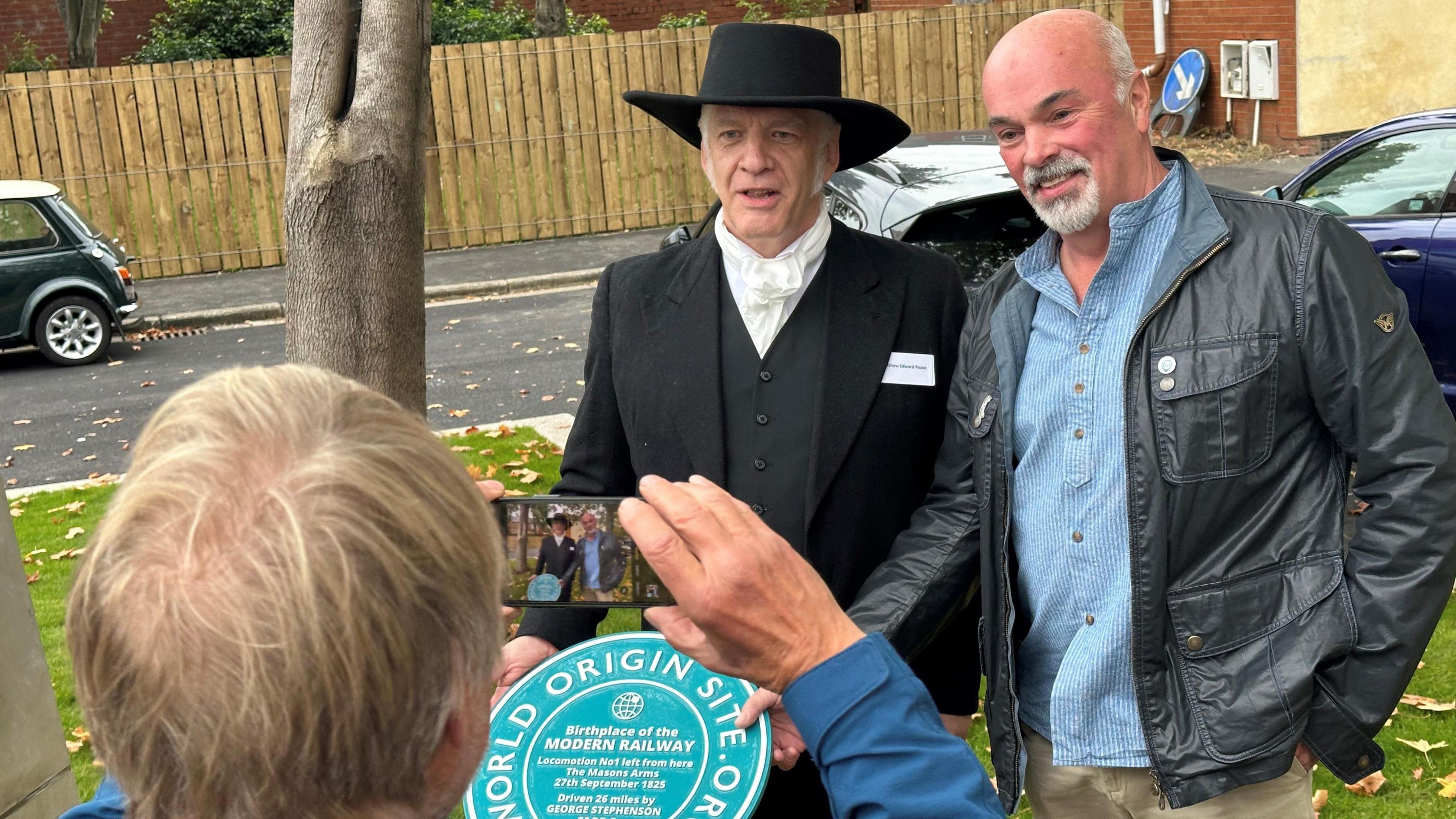 A man takes a picture on a phone of Matthew Edward Pease and another man. Matthew is wearing a black coat and waistcoat with white cravat and wide brimmed black hat. In front of them is a round green plaque marking the spot where the original engine left from. 