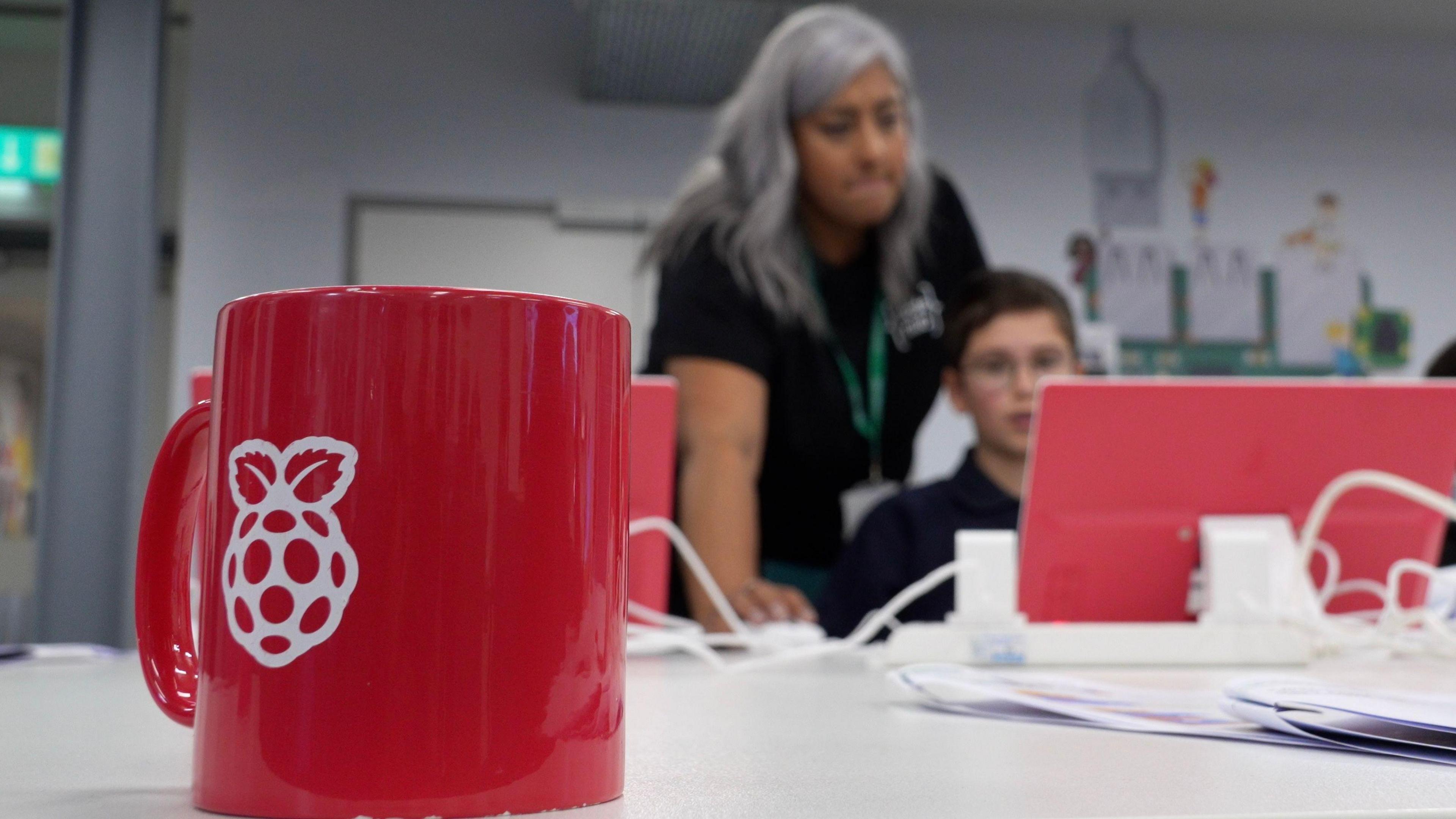 A red Raspberry Pi-branded mug sits in the foreground of a tech-focused workspace. Behind it, two (an adult and a child) are engaged with a computer screen. The adult has long grey hair and is helping the child with their work.