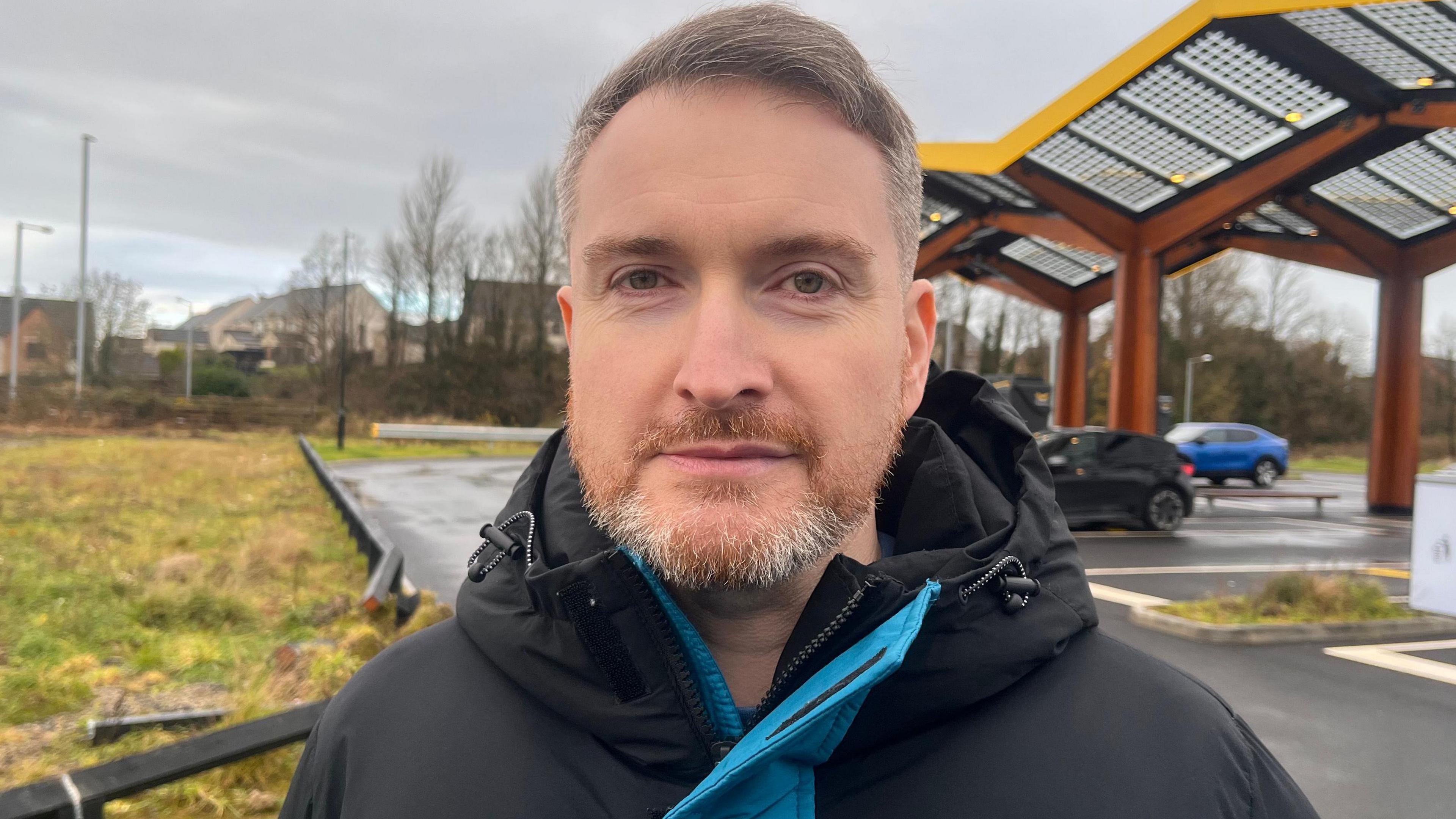 Shane Hagan is outside. He's wearing a black and blue coat. There are car in the background.