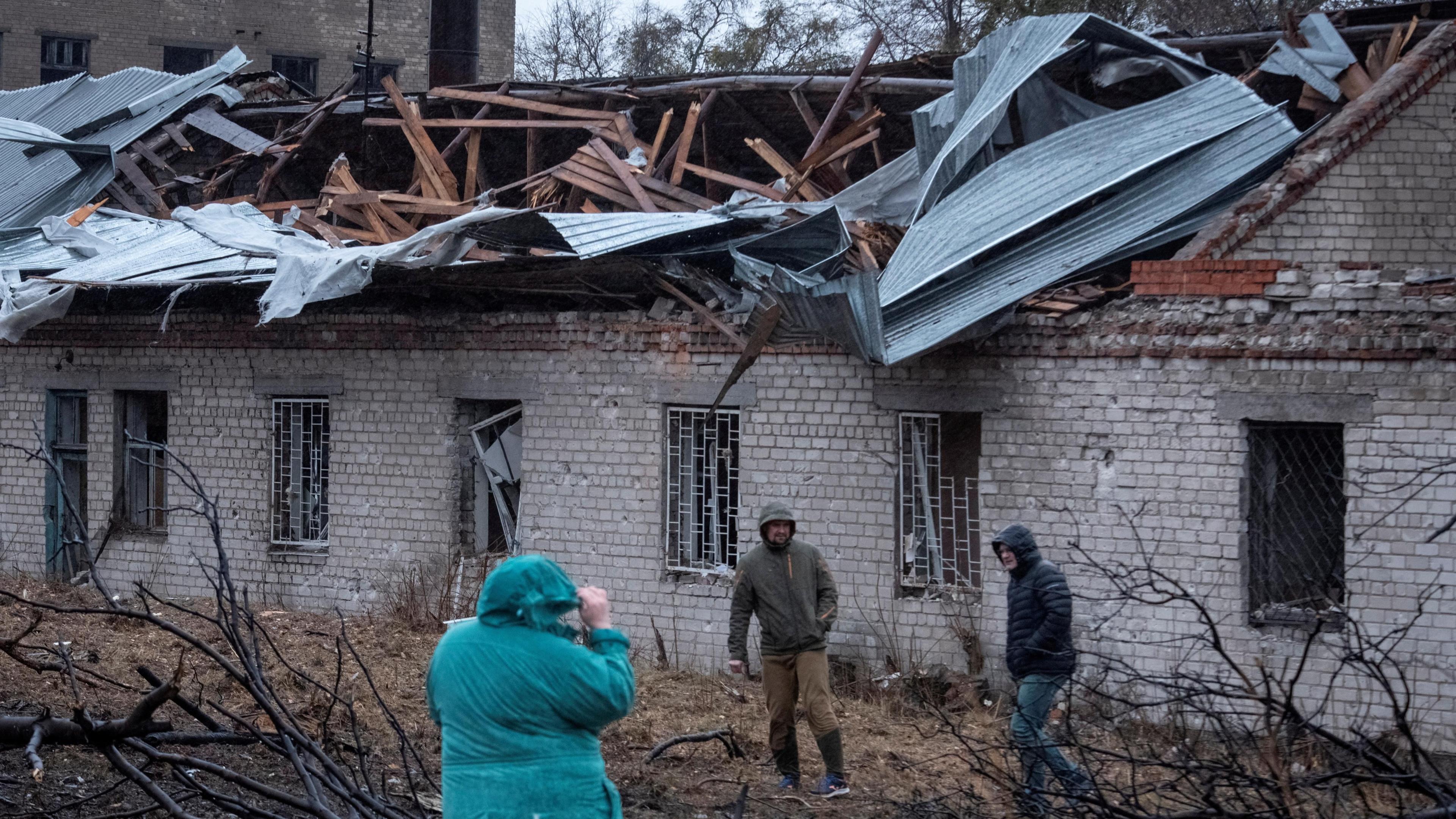 A woman and two men wearing coats with the hoods up stand in front of a one-story building with a destroyed roof and windows