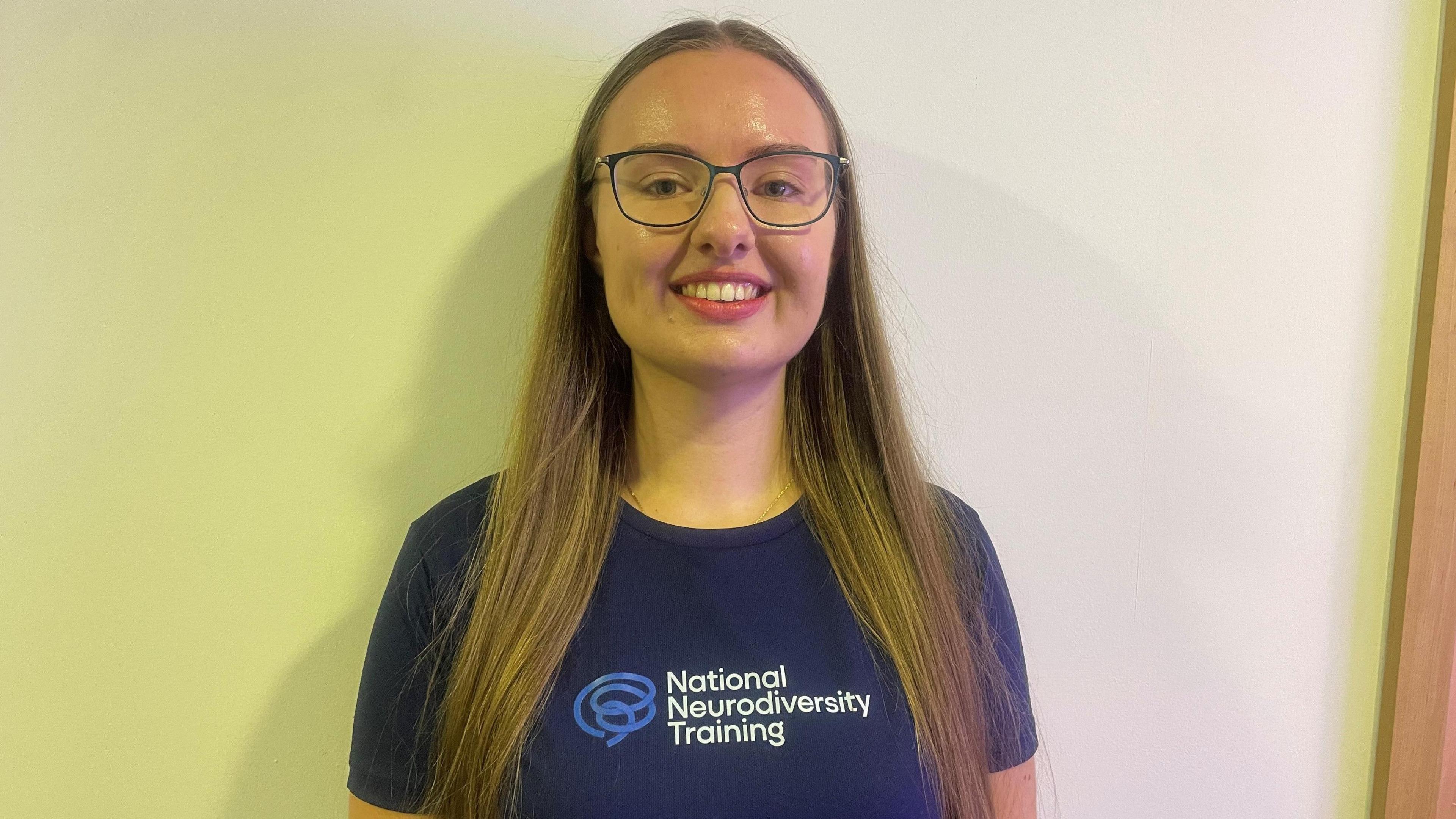 A woman with glasses and long brown hair is in front of a yellow and white background. She is wearing a blue T-shirt including the words National Neurodiversity Training.