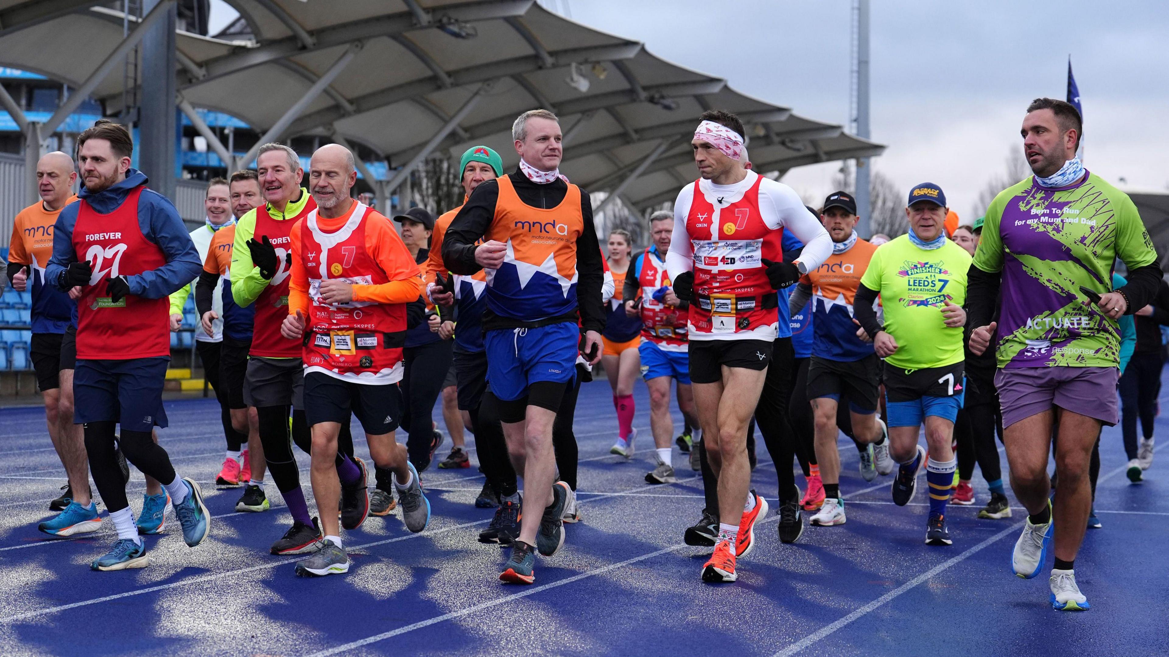 A group of about 20 people in brightly-coloured running gear run alongside and behind Kevin Sinfield on a blue running track with a spectator stand in the background.
