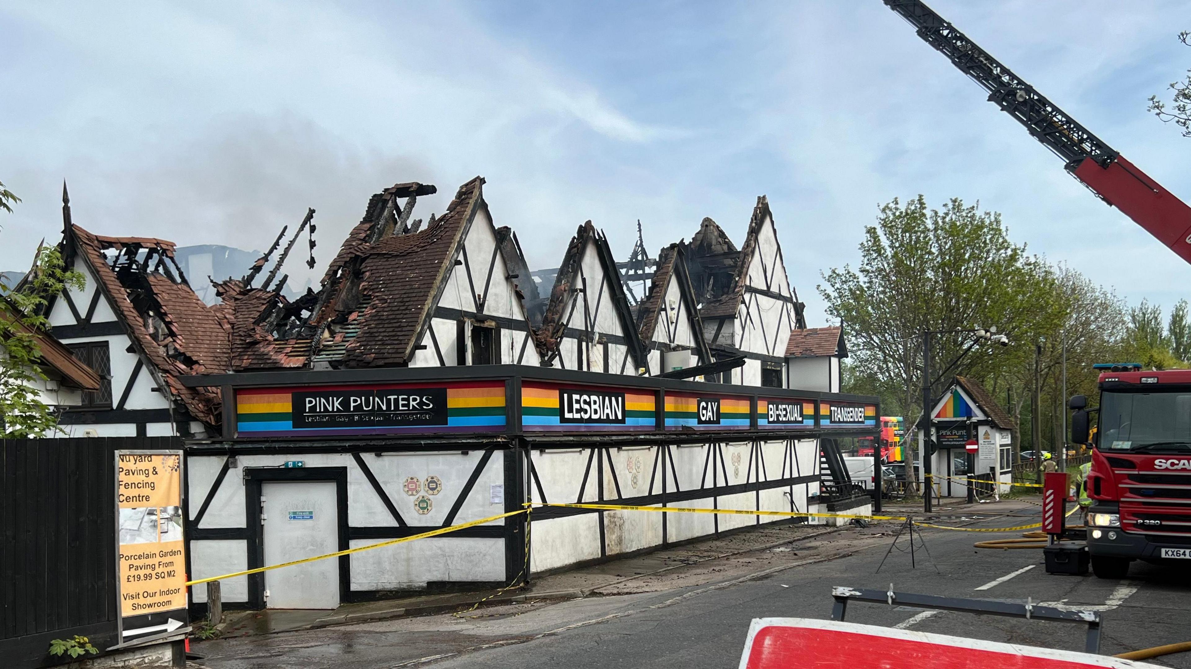 A nightclub, pictured in the middle of the day, with a roof that has been destroyed in a fire. A fire engine is parked opposite the venue. There is a long aerial ladder rising into the sky above the building. Some smoke can be seen billowing out of the venue.