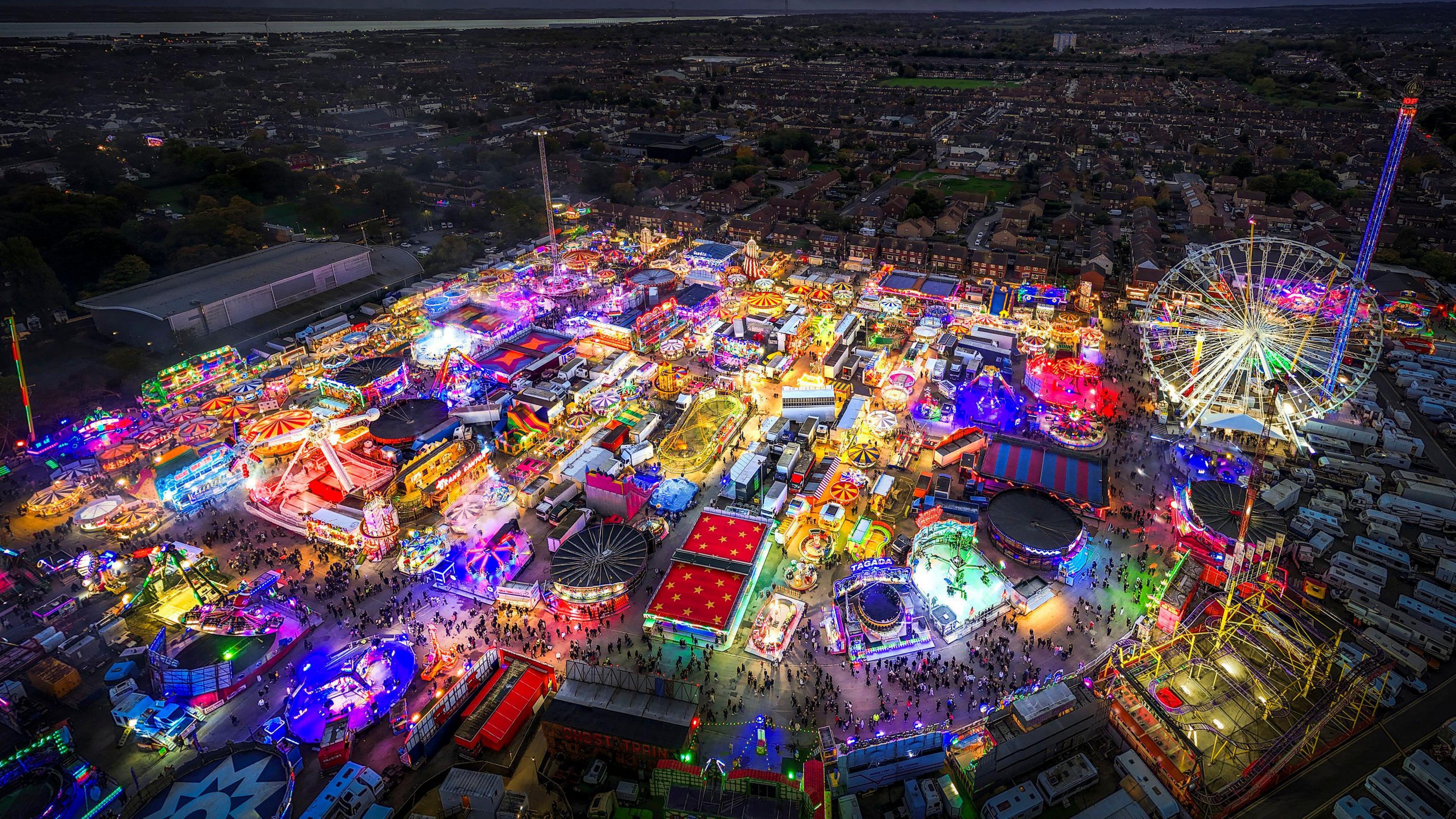 An aerial view of a funfair with an adornment of neon-coloured lights surrounding a city at night. You can see a Ferris wheel, tall pole-looking rides, swirls and stars.