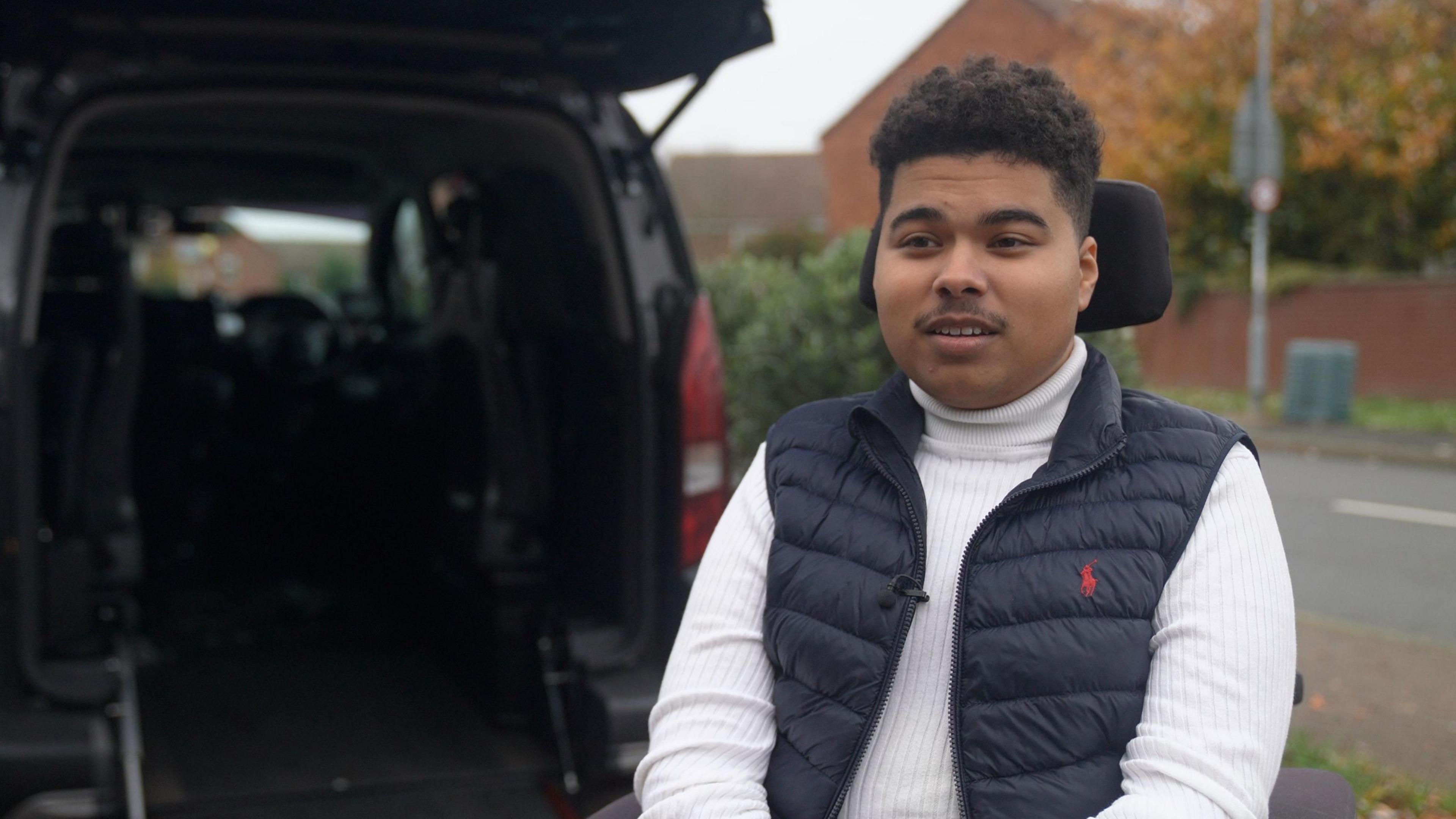 21 year old man, Maxwell, sitting in a wheelchair in front of the open boot of his wheelchair-adapted van, with its ramp