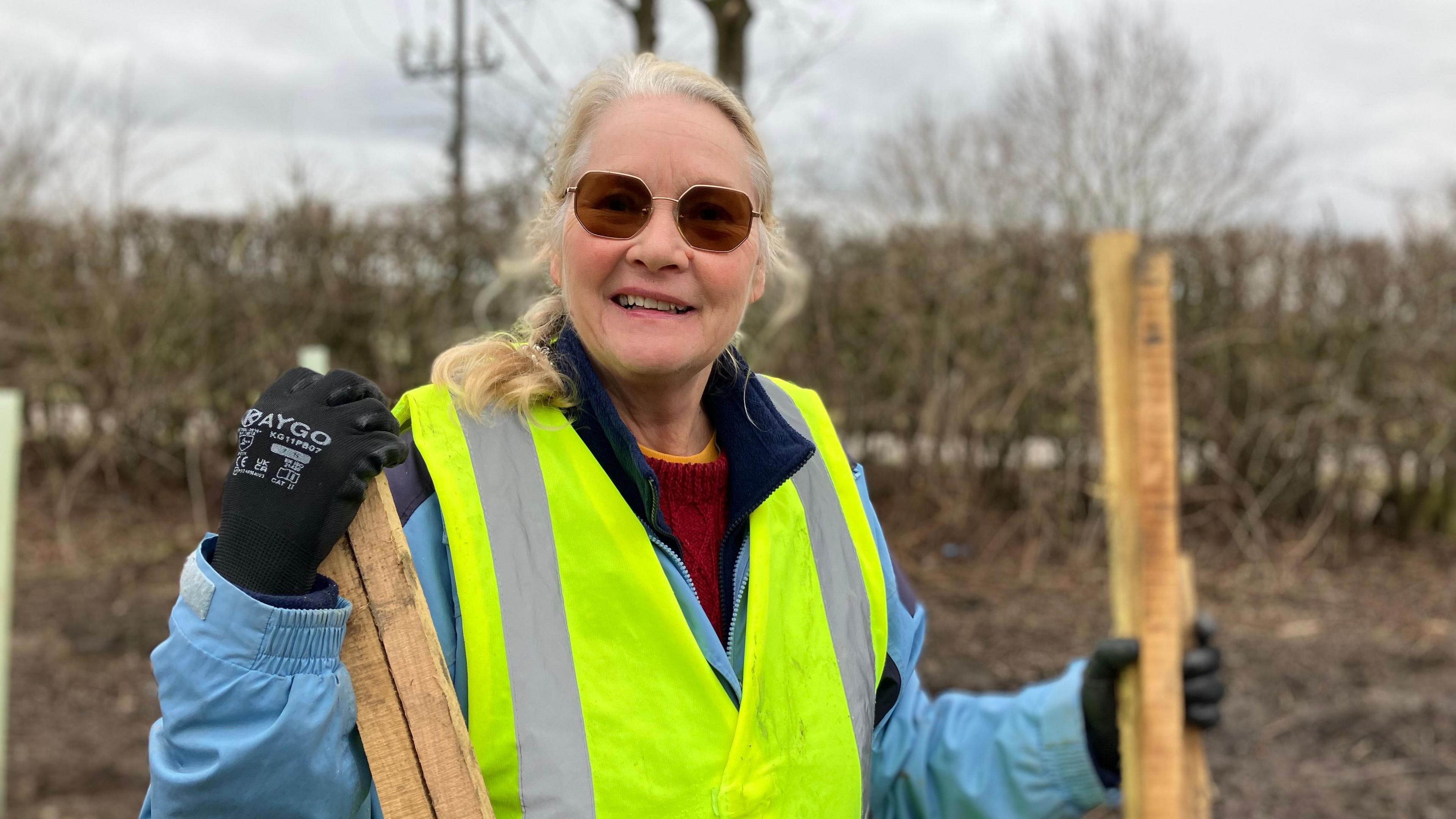 A woman smiling, wearing a reflective vest. She has long hair and is wearing glasses. A hedgerow can be seen in the background. She is wearing gloves and holding long wooden poles. 