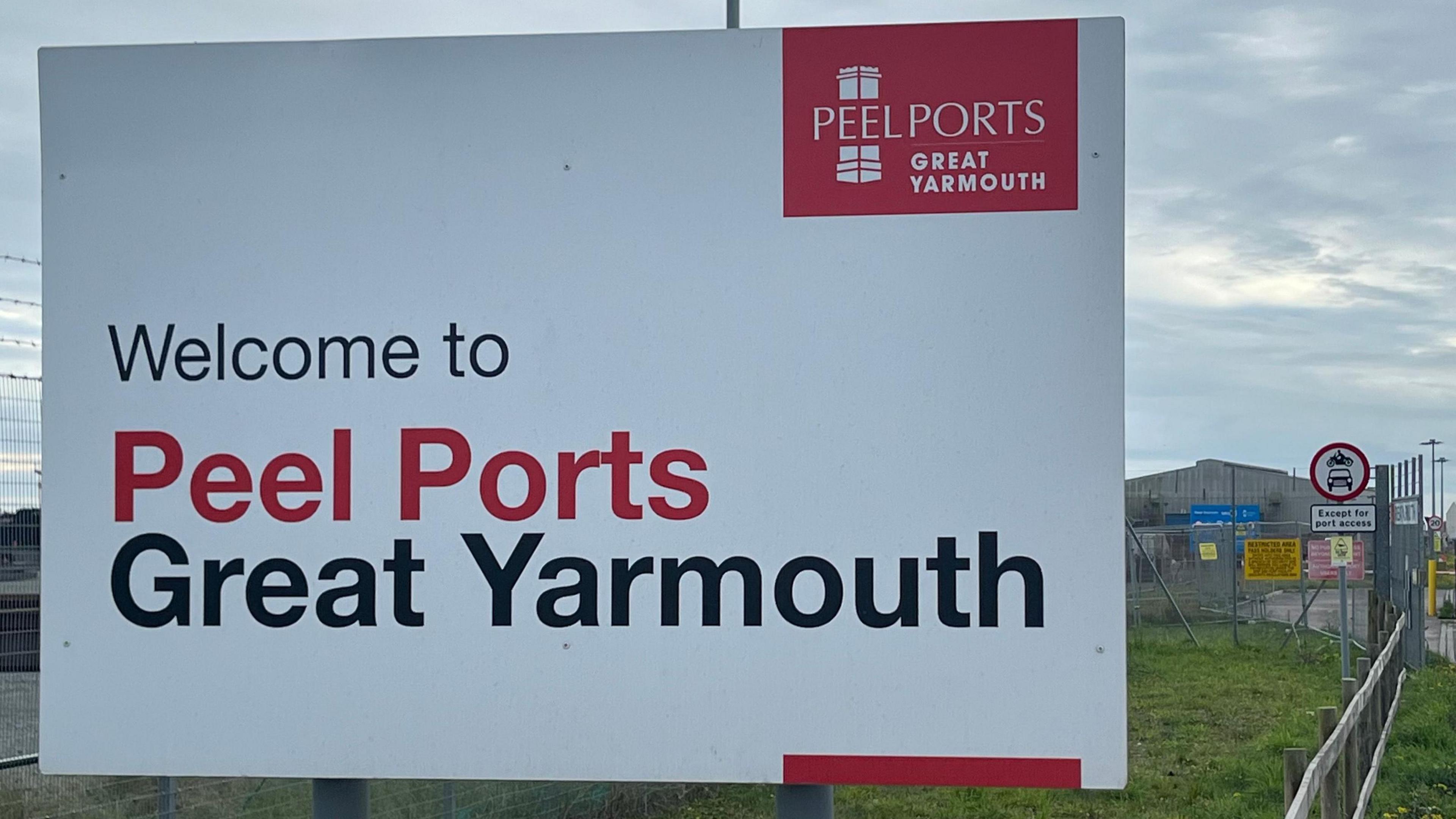 An entrance sign into the Port of Great Yarmouth in Norfolk. Is shows a white sign with red and black letters. An entrance gate can be seen in the distance.