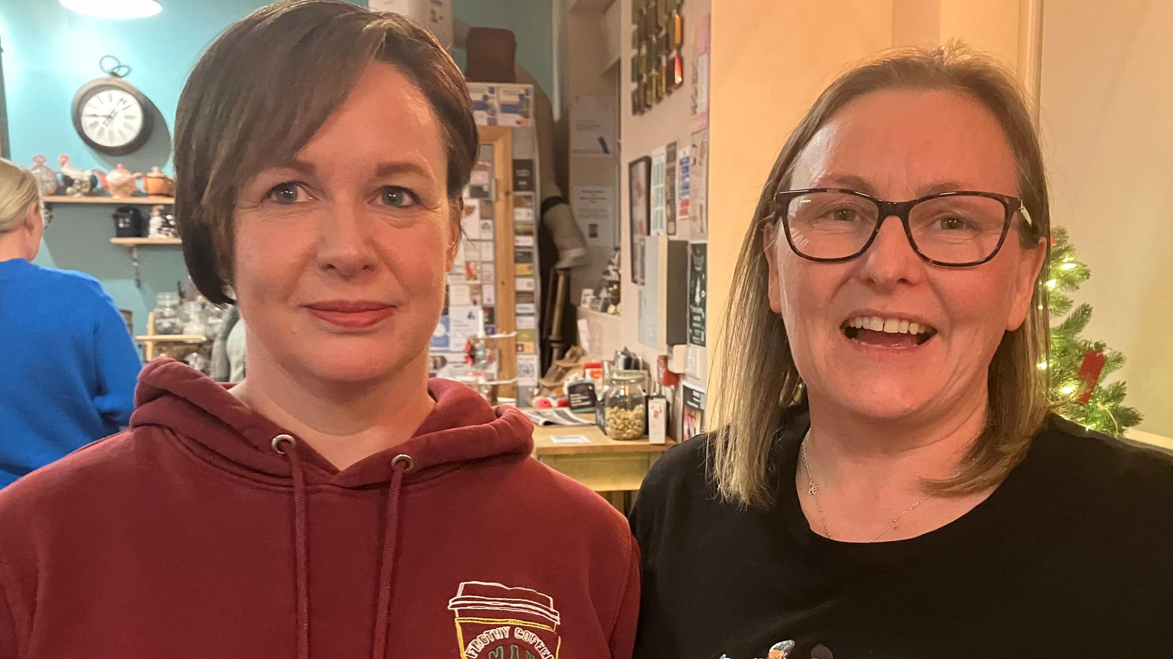 A picture of two women. On the left is a woman with short dark hair, parted on one side, who is wearing a dark red fleece. On the right is a woman with shoulder length light brown hair, wearing a black t-shirt with a giraffe on it.