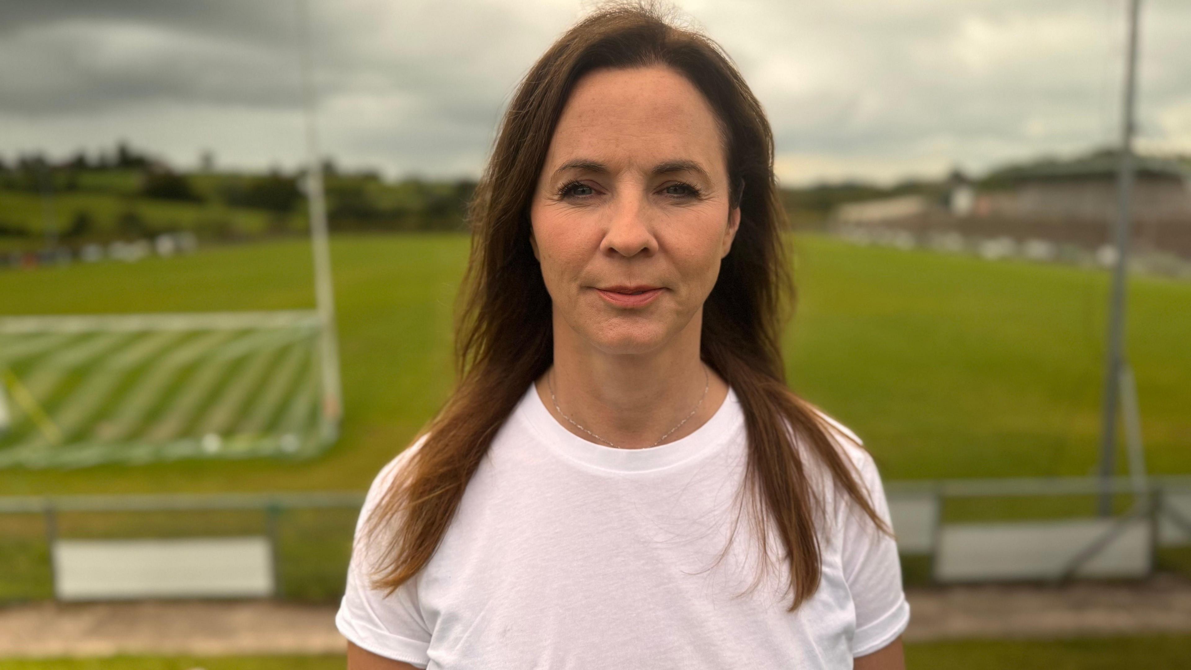 A woman is standing with a sport filed behind her. She has brown hair and is wearing a white top.