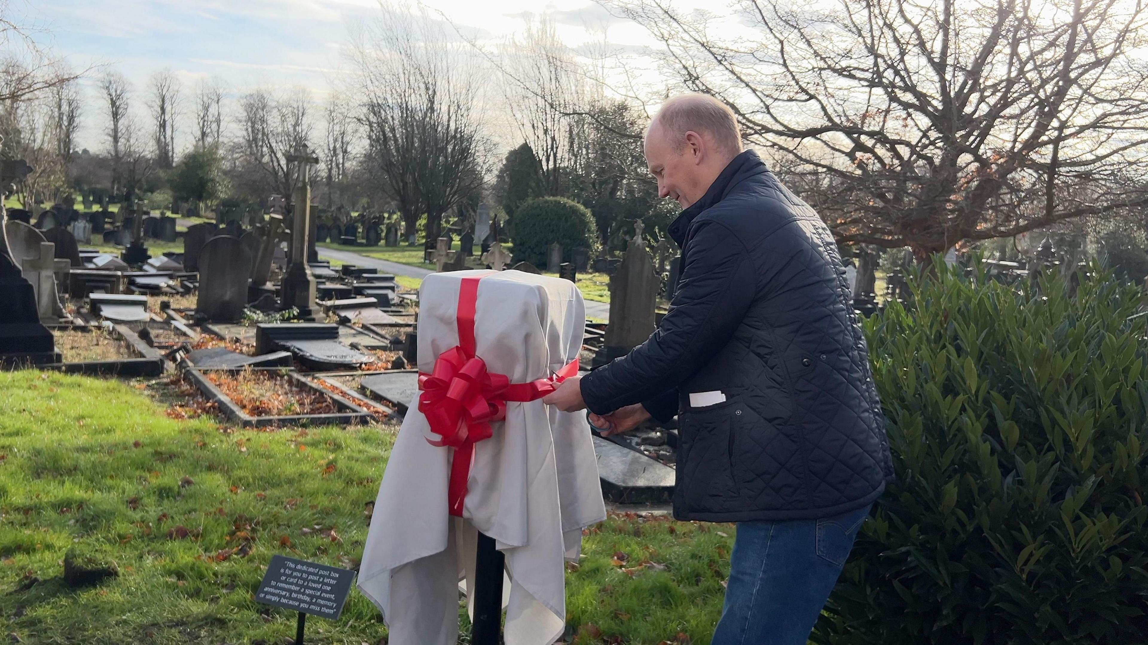 A man cutting a red ribbon to unveil a 'Letters to Heaven' post box at Nottingham Road Cemetery.