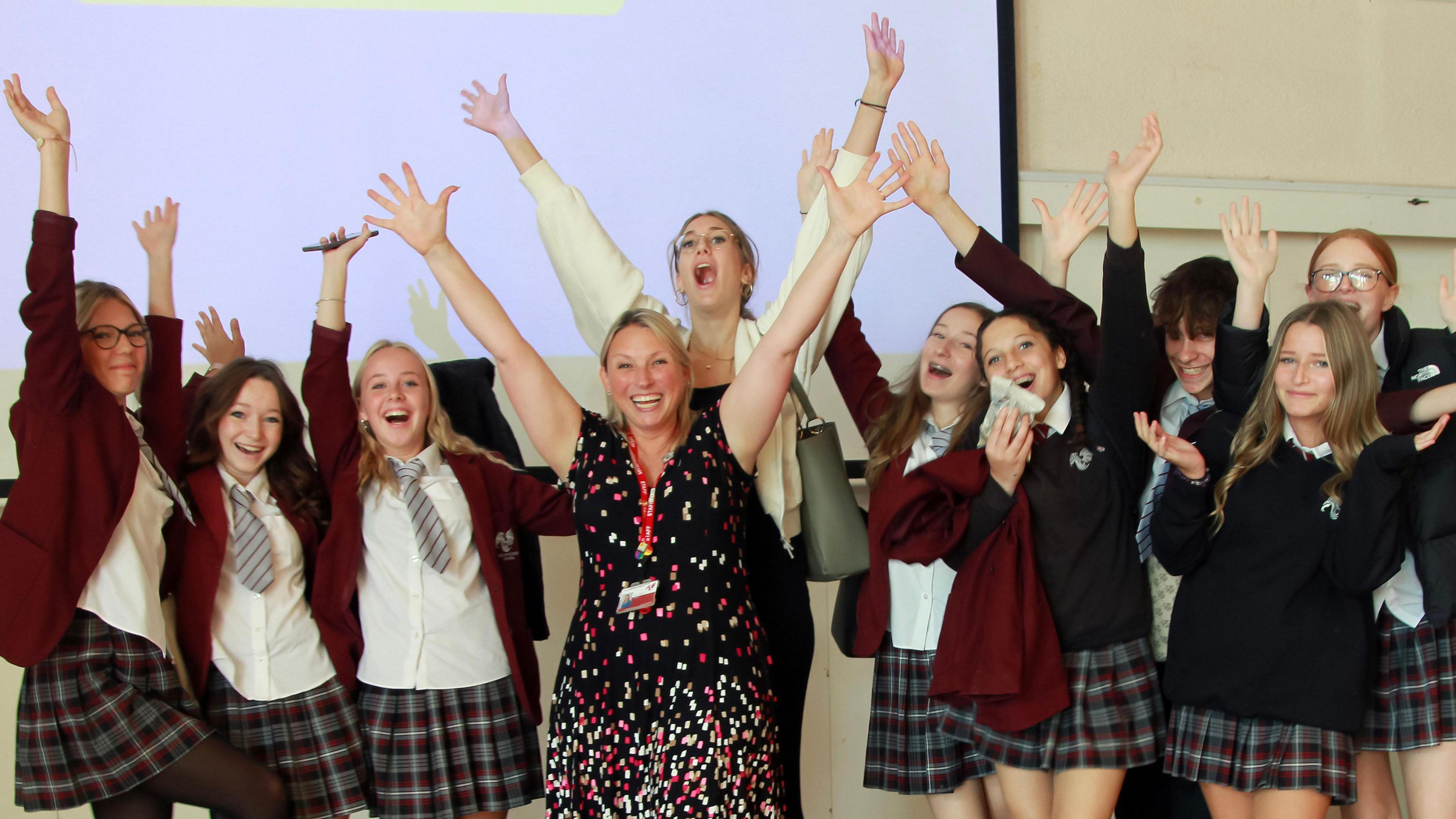 A group of school students around Ms Watson. All have their hands in the air and are celebrating.