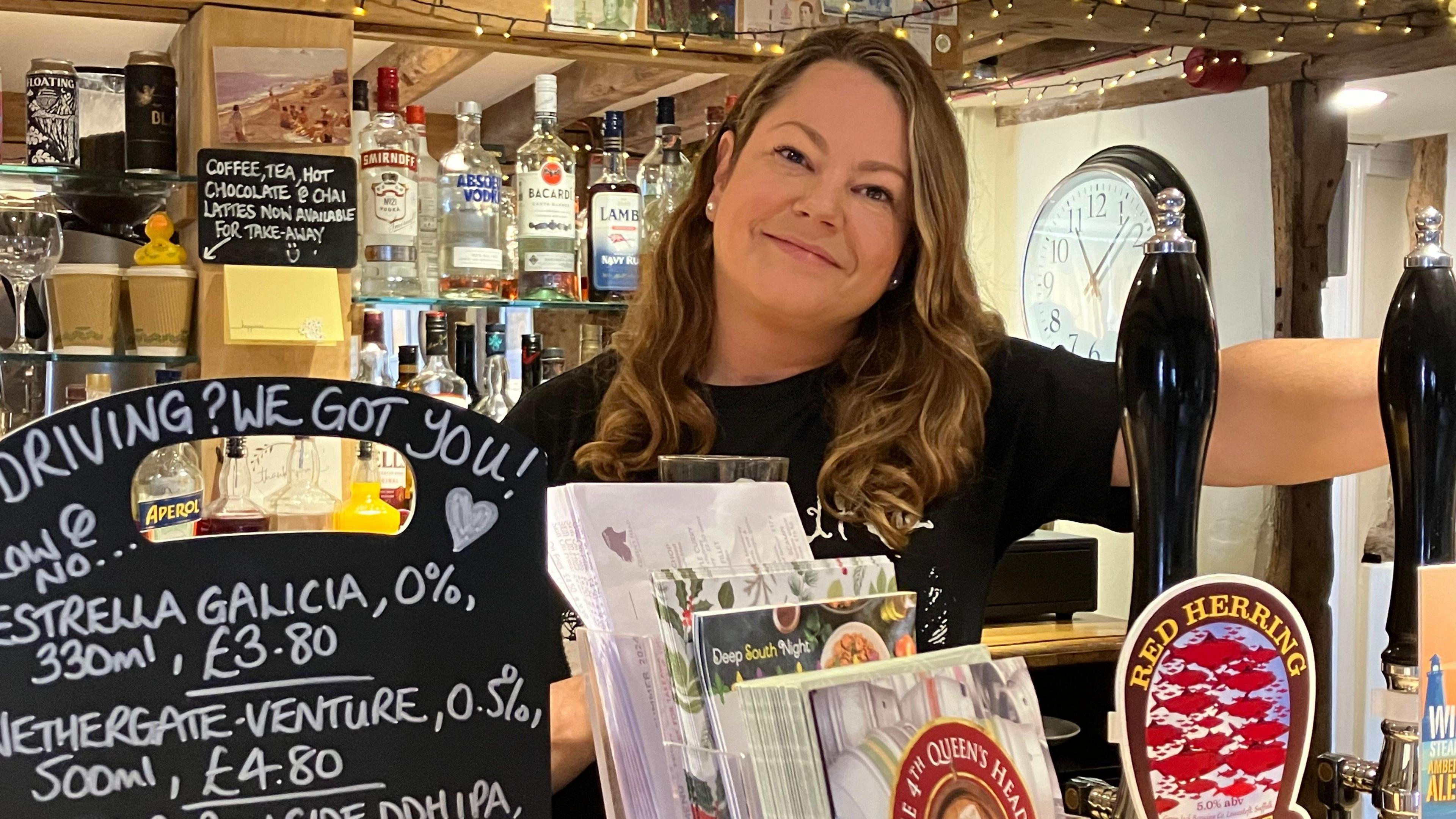 Person wearing black shirt stands behind a bar covered with leaflets and a small blackboard