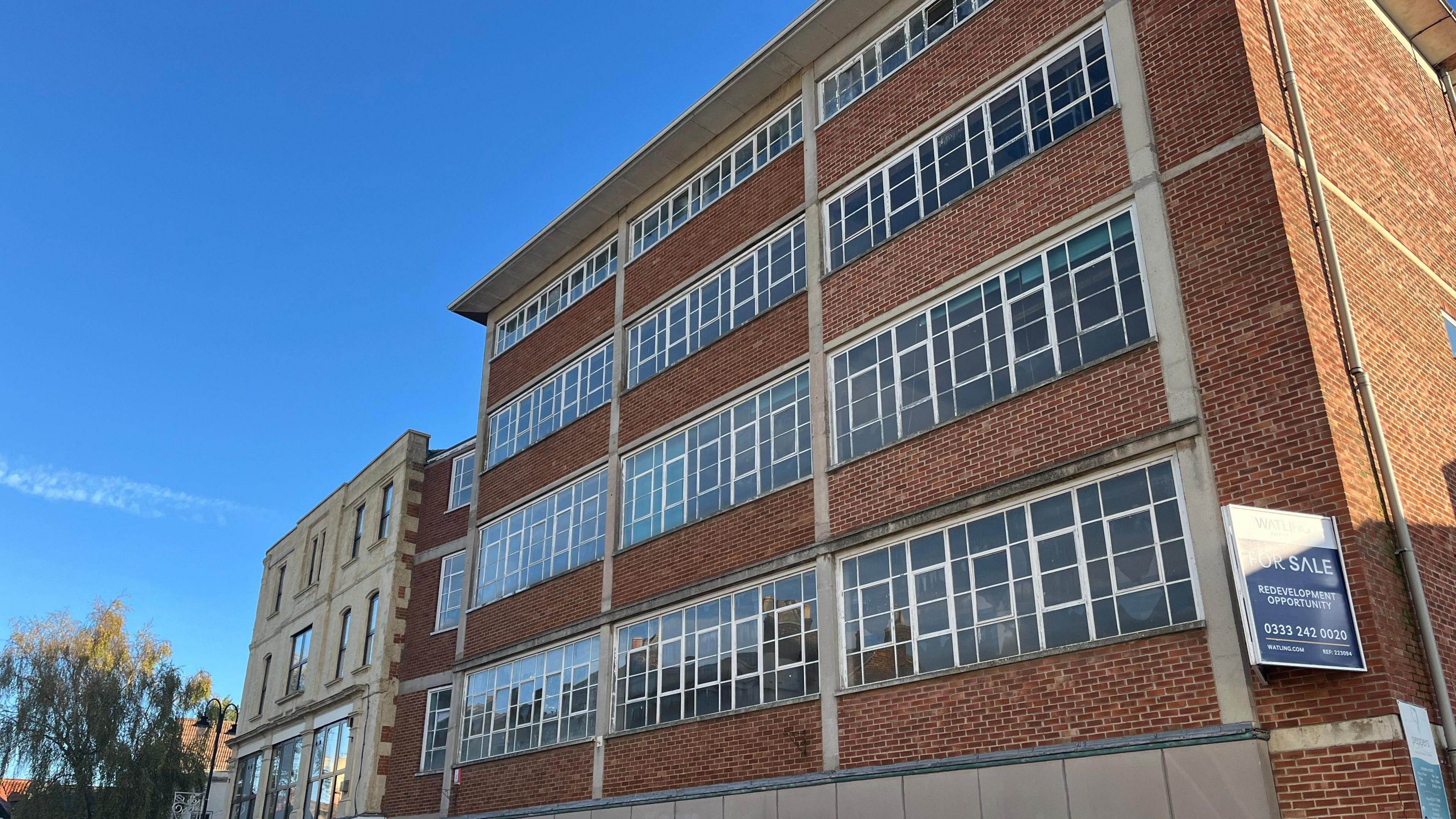Red-brick building about four storeys high with a 'for sale' sign on the right. It also has large windows with white frames