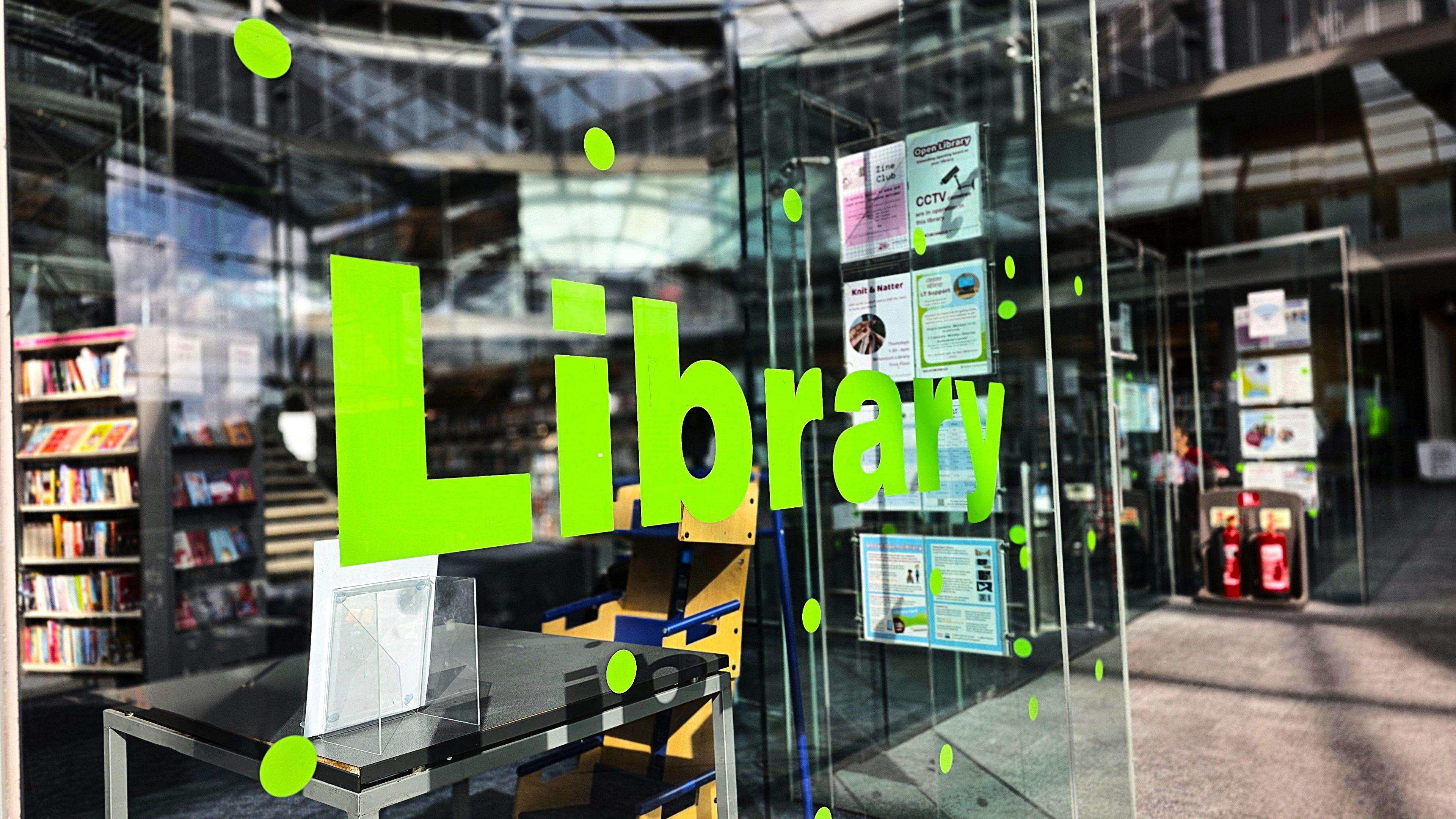 The front of the first floor of the Norfolk and Norwich Millennium Library which is located in The Forum in Norwich city centre. The building is made of glass and there is green lettering on the panel which says Library. Behind the panel is rows of books on shelves.