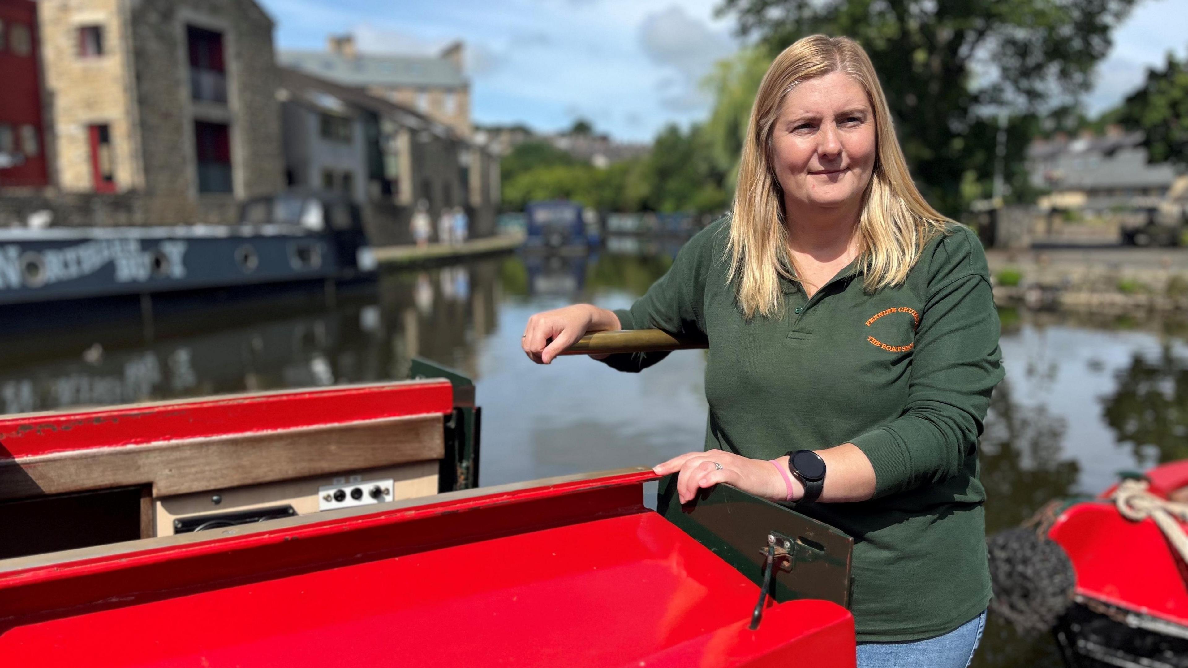 A woman with long blonde hair stands at the back of a red narrow boat. She is holding onto a tiller and you can see the canal and another narrow boat in the distance. It is sunny.