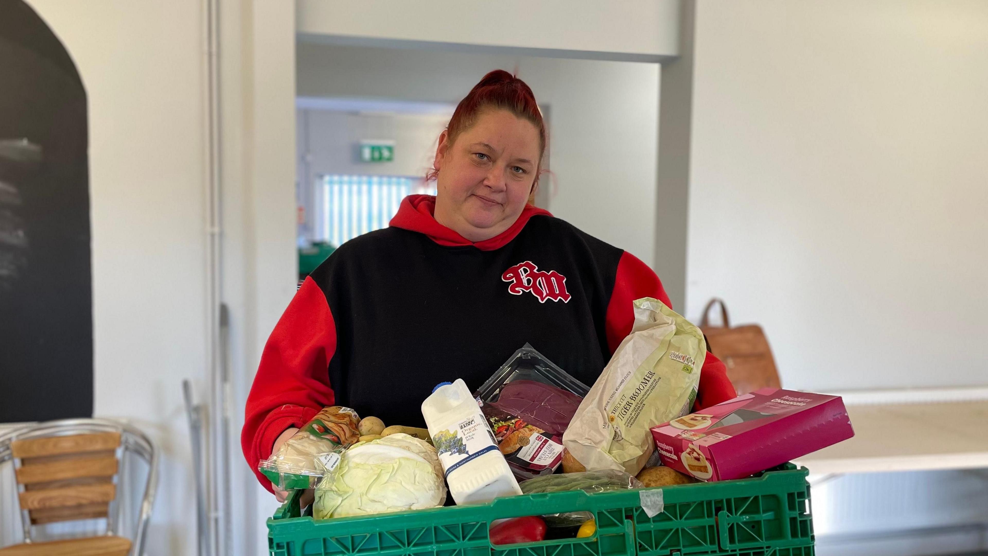 Gemma Douglas is holding a green plastic box full of food, with bread, cabbage, steaks, milk and a box of tarts.
