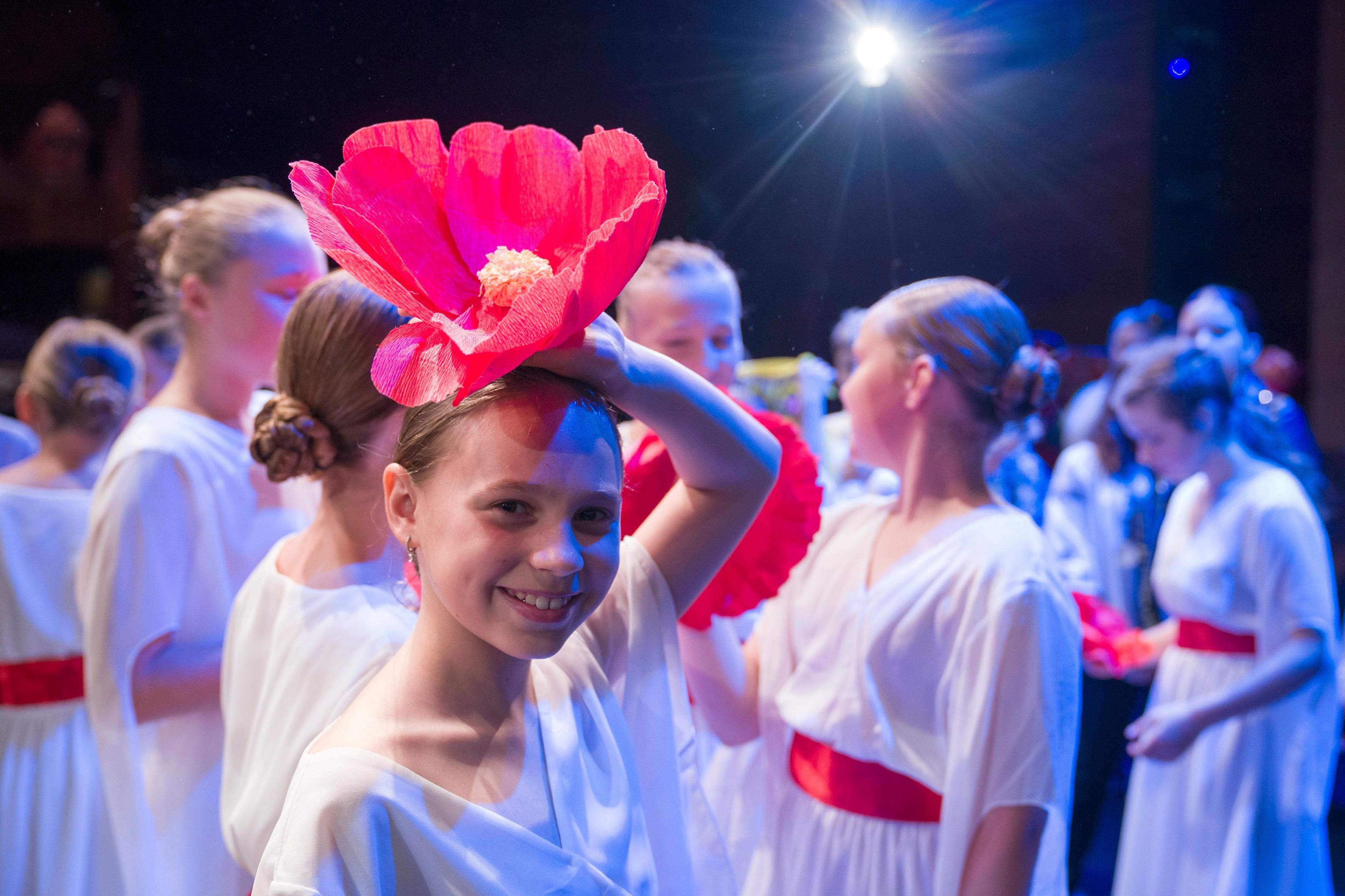 A young Ukrainian girl in a white dress smiles and holds a big red flower over her head while on stage. Similarly-dressed children stand in a group behind her under the theatre lights.