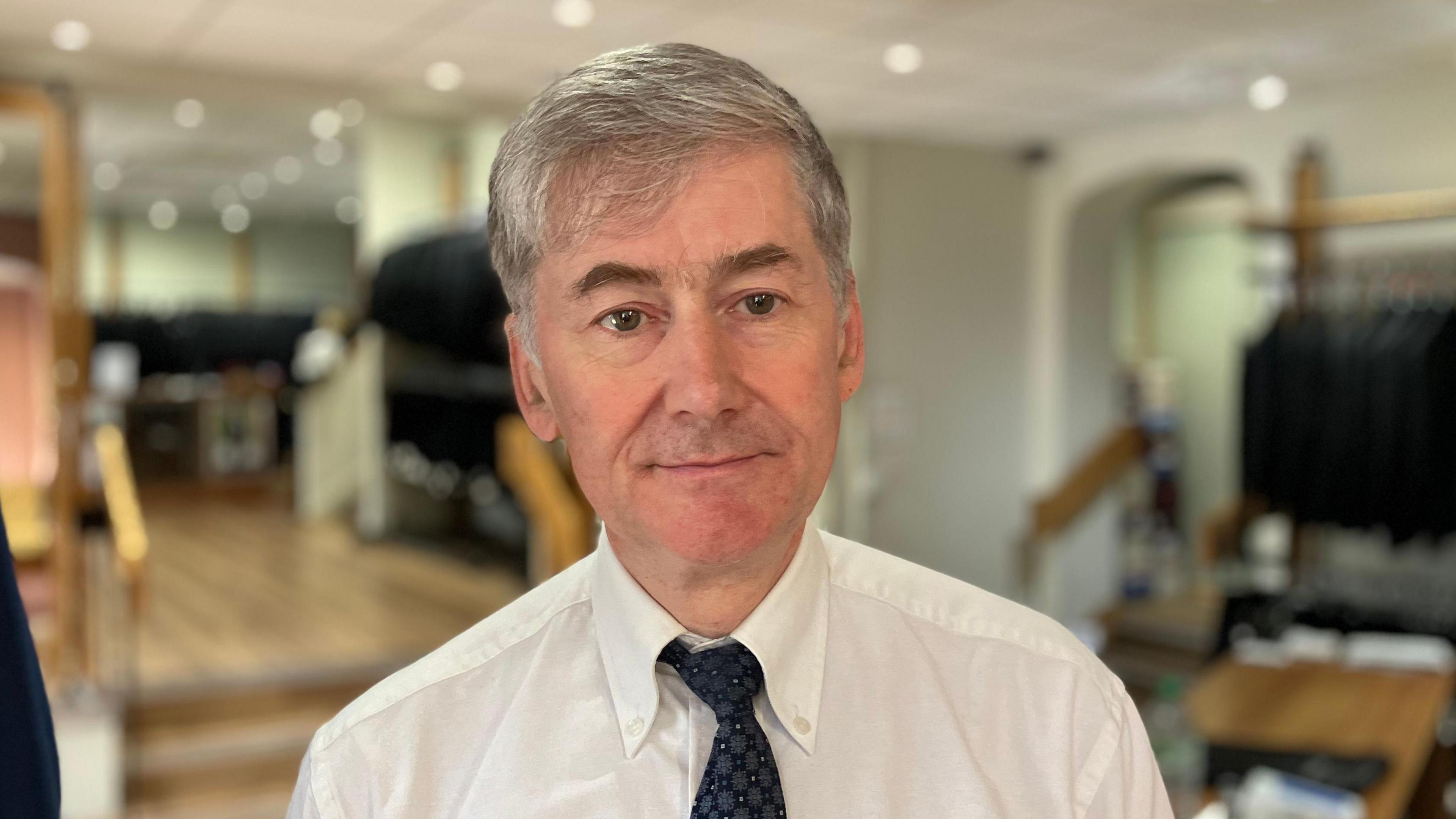 Neil Warwick, standing in a shop. He has grey hair and is wearing a white shirt, blue tie and a white shirt with an oxford collar.