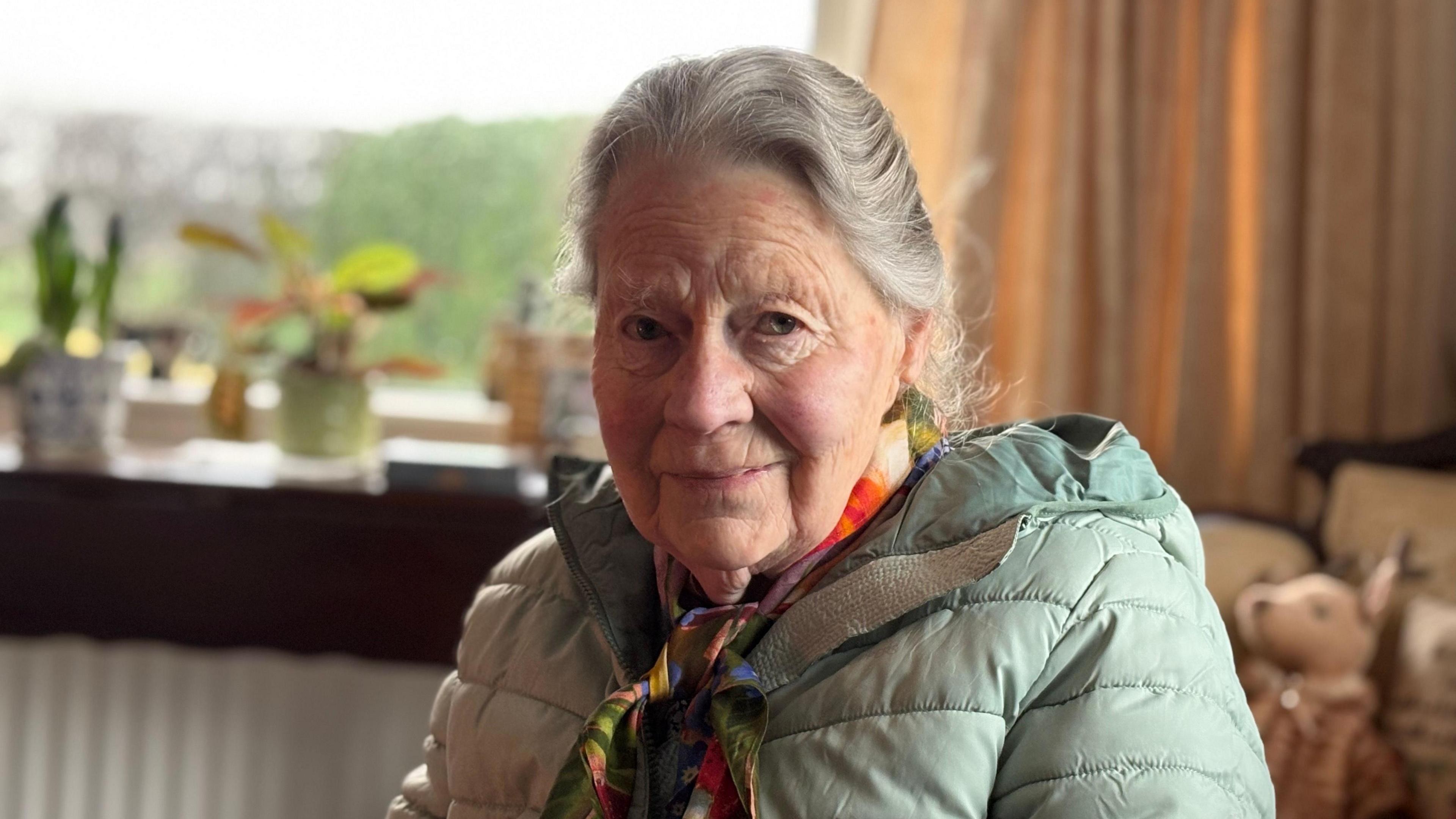 Elinor Farquhar. She has grey hair tied back, wearing a mint green puffer coat and multi-coloured silk scarf.