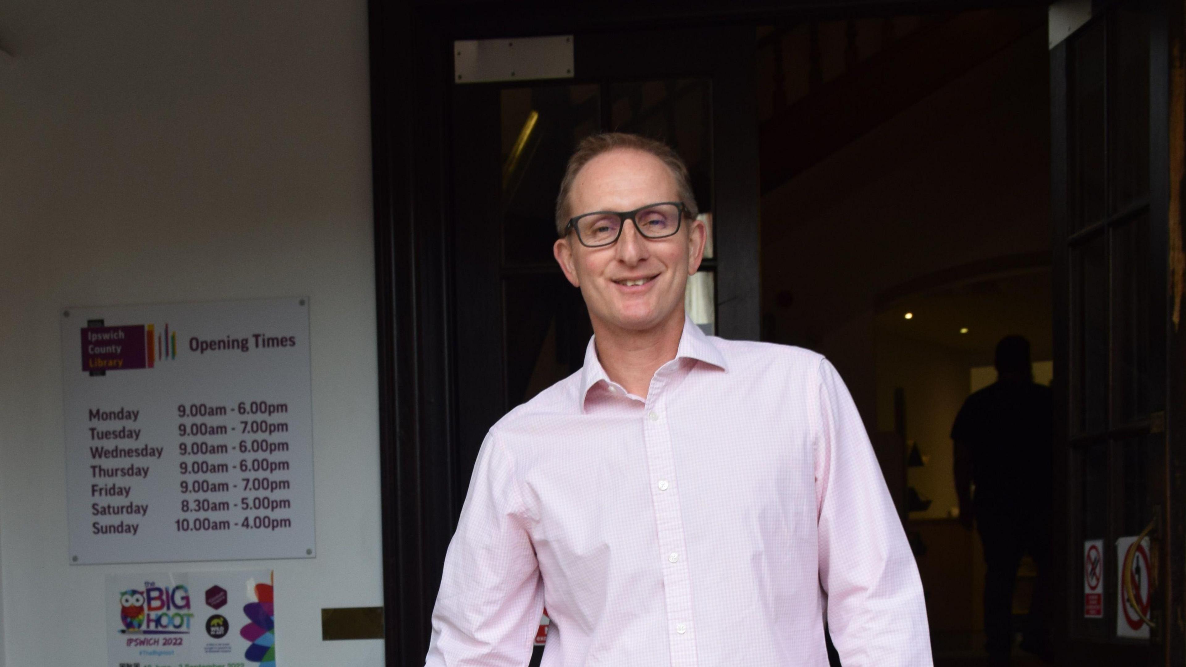 Bruce Leeke is standing in front of a library and smiling. He is wearing a pink shirt and glasses, with receding light brown hair.