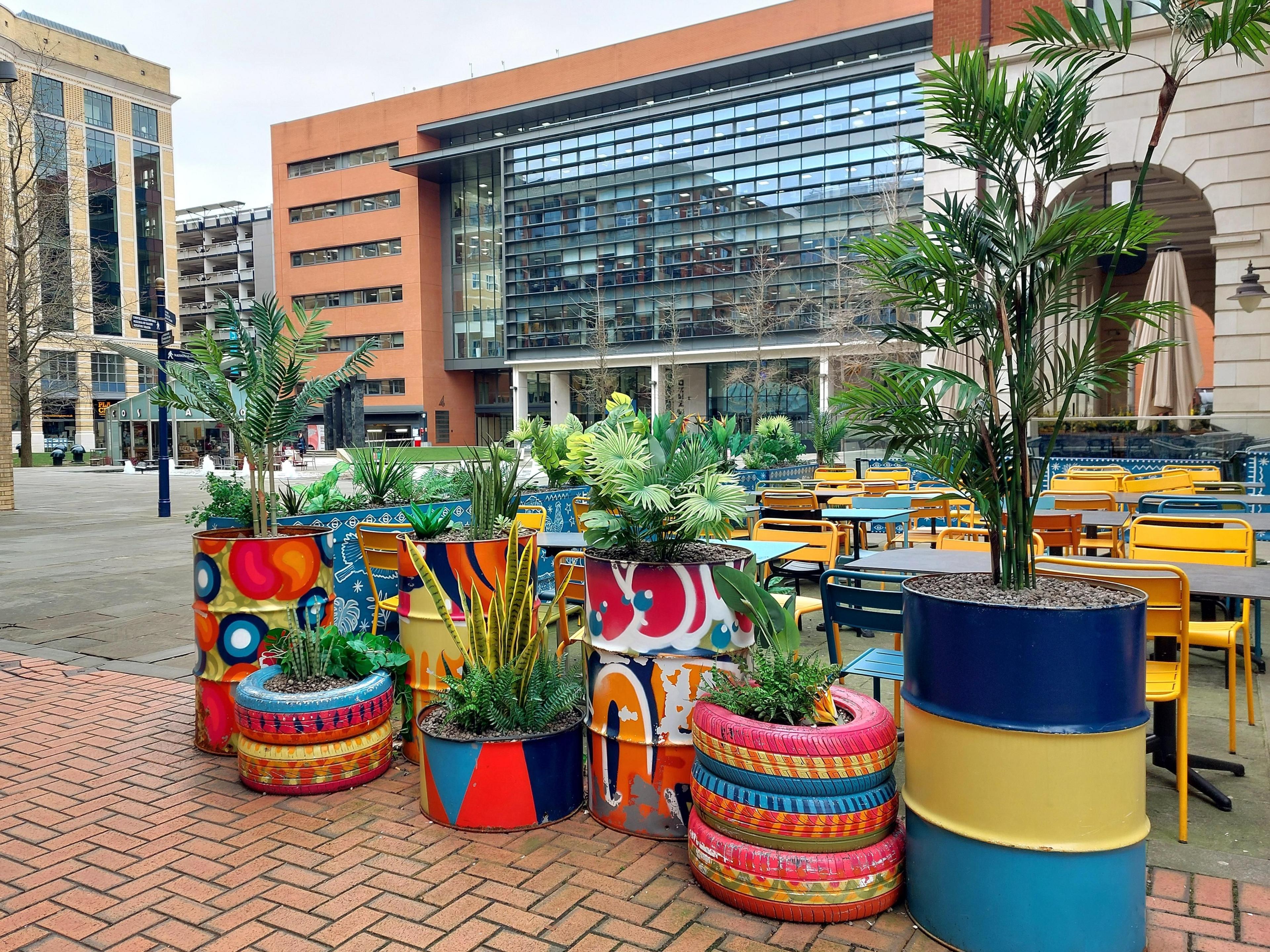 A number of planters made from oil drums and stacks of tyres, all painted in vibrant colourful patterns with ferns and succulents planted in them. Behind, outdoor cafe seating and office buildings are visible. 