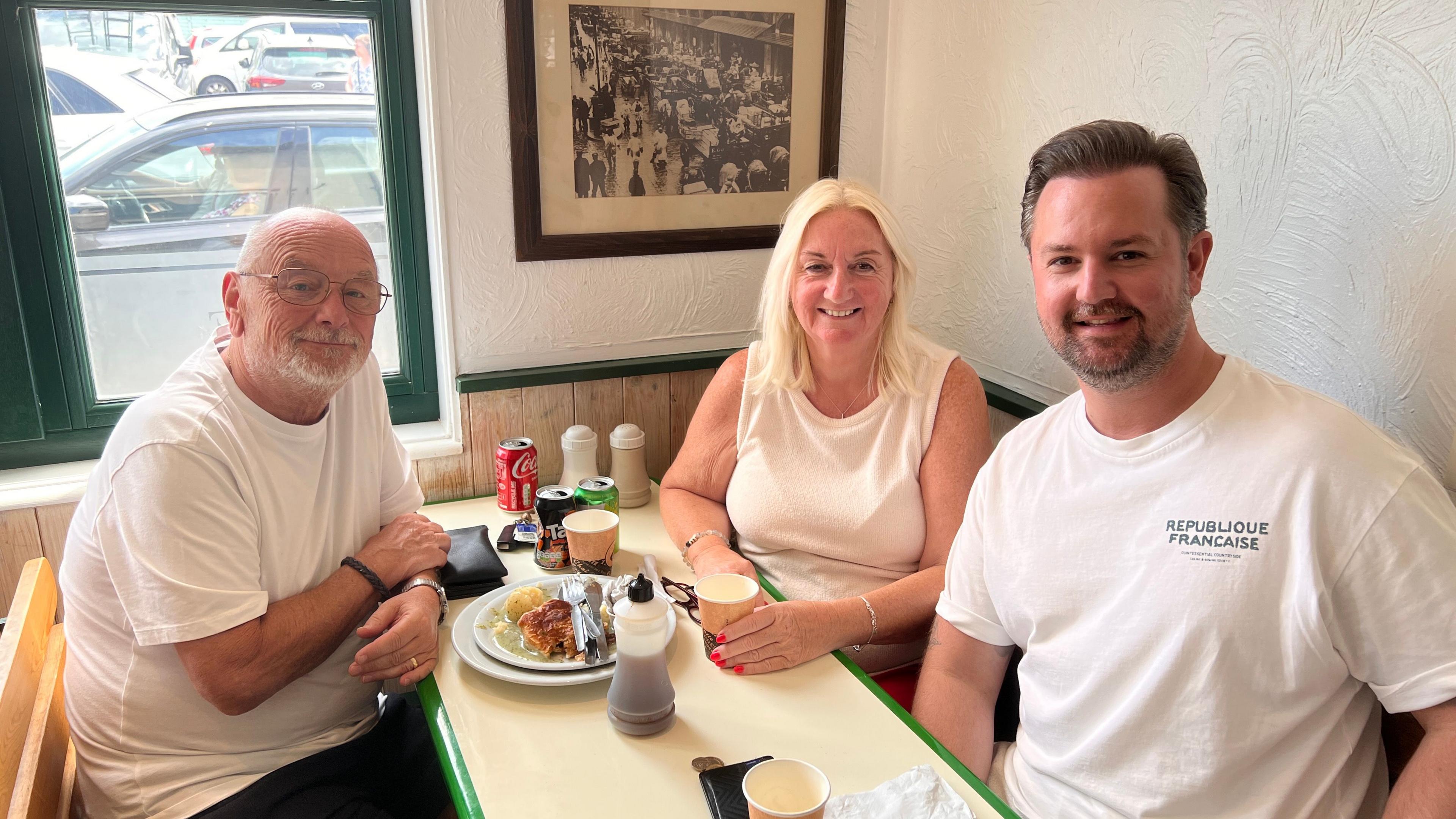 A family at a dining table in a cafe. There are finished plates stacked with pie and mash. A bald man is sitting with his arms on each other wearing a white t-shirt and smiling at the camera. Opposite is a lady in a white top with white shoulder length hair next to a man in his late thirties both are smiling at the camera.