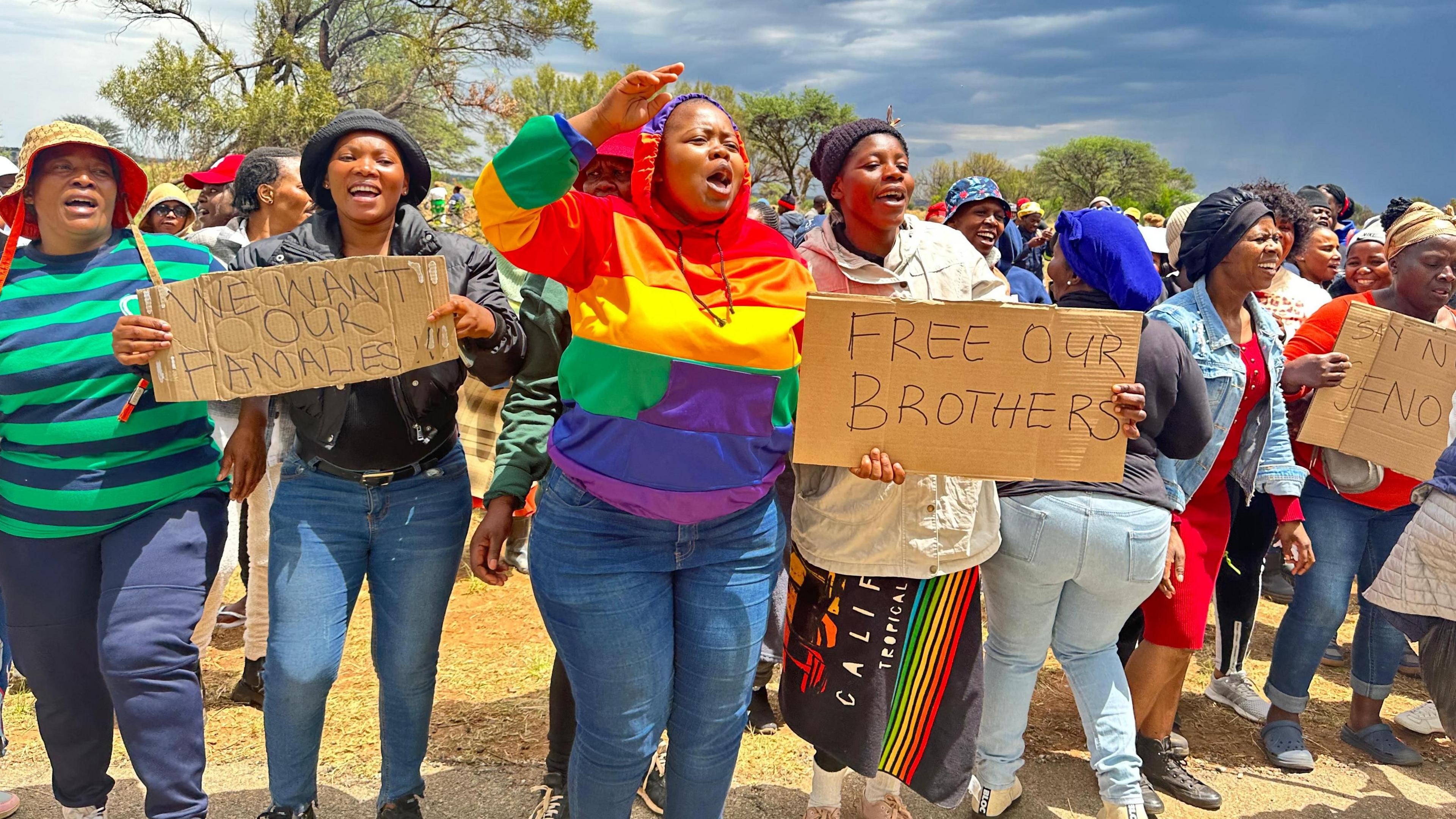 A group of female protesters, with one dressed in a rainbow hoodie, are shouting. They are holding placards saying - "free our brothers" and "we want our families"