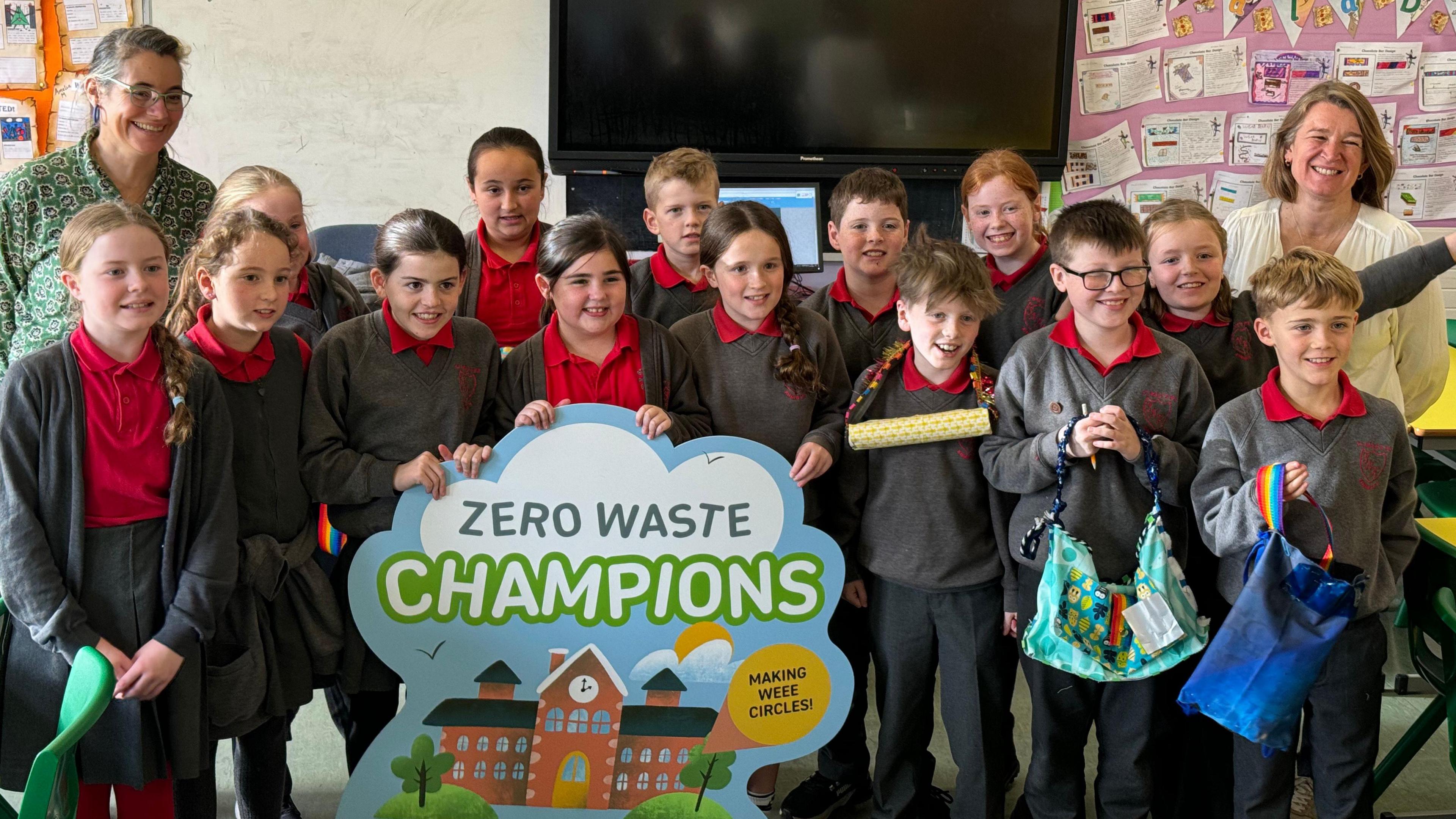 Fourteen pupils wearing red and grey uniforms hold some of their recycled creations while holding a large colourful sign saying 'Zero Waste Champions'. They are stood in the middle of a classroom with teachers on either side.