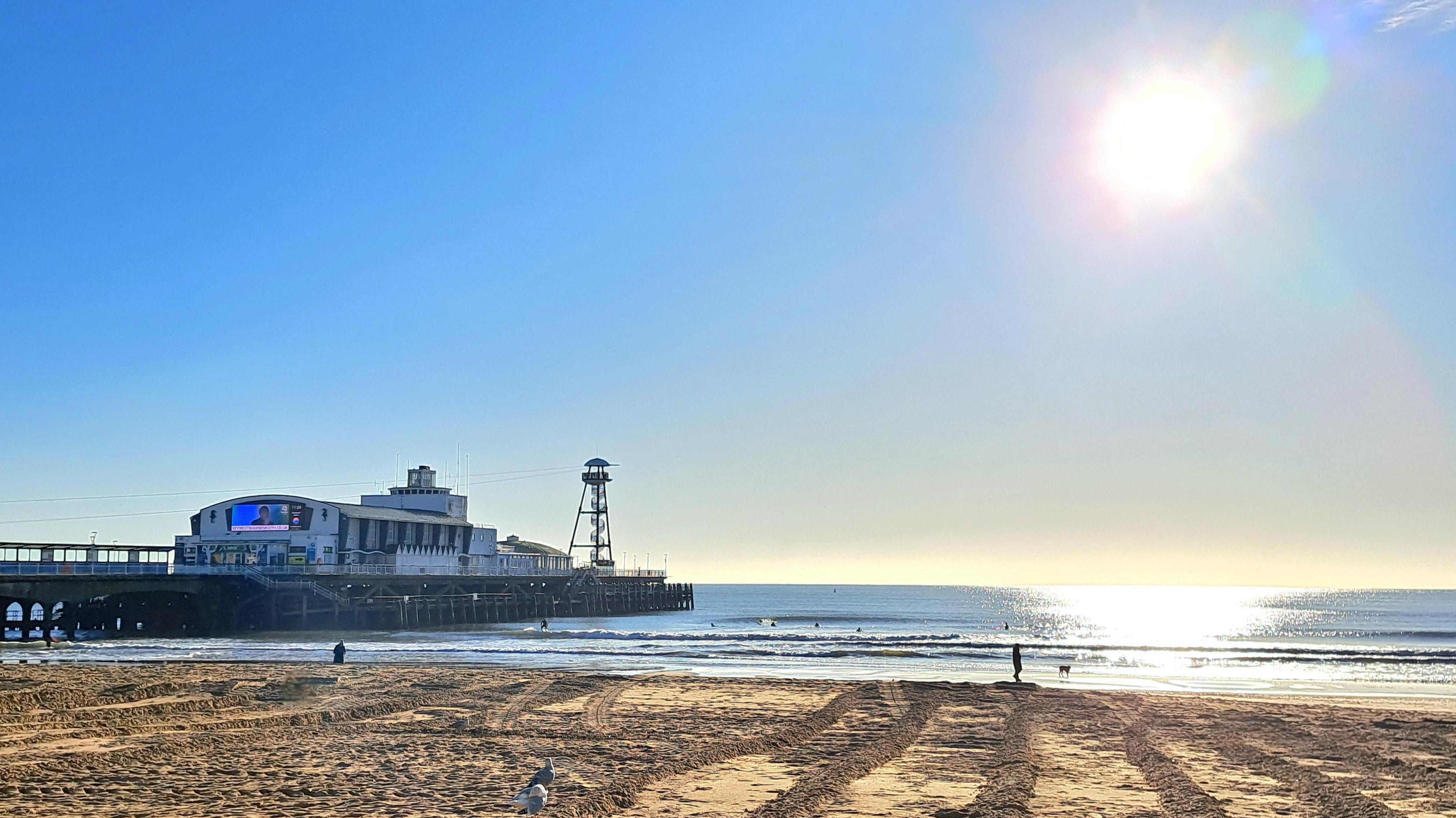 A sandy beach on a sunny day. Bounemouth Pier stretches out into the sea and you can see surfers in the water. The sky is glowing in the sky and reflecting in the water. You can see two walkers, a dog and several seagulls on the sand. 