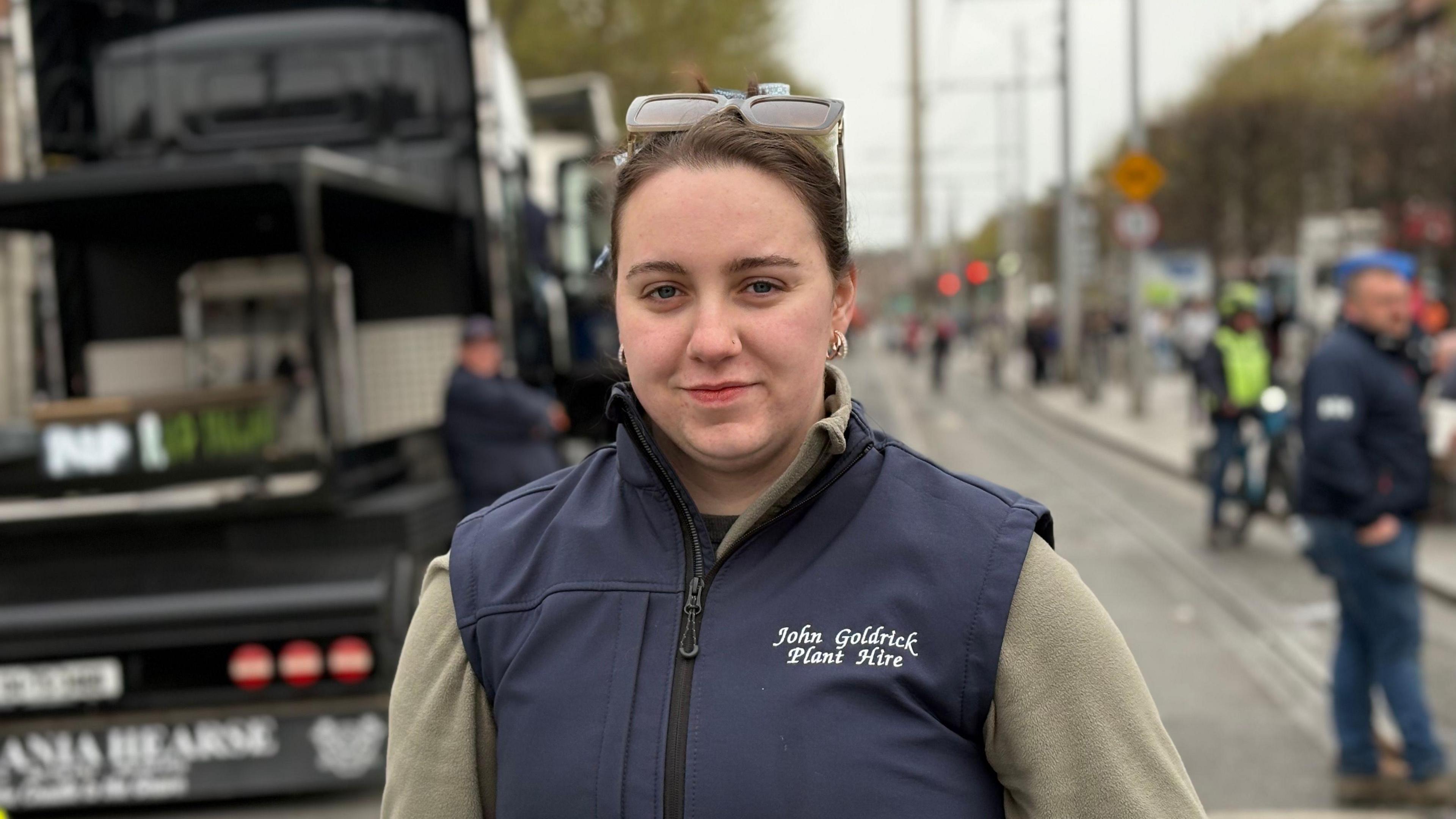 A woman with brown hair smiles at the camera. She is wearing a grey jumper and a dark-blue gilet-jacket. Behind her is a busy urban street. 