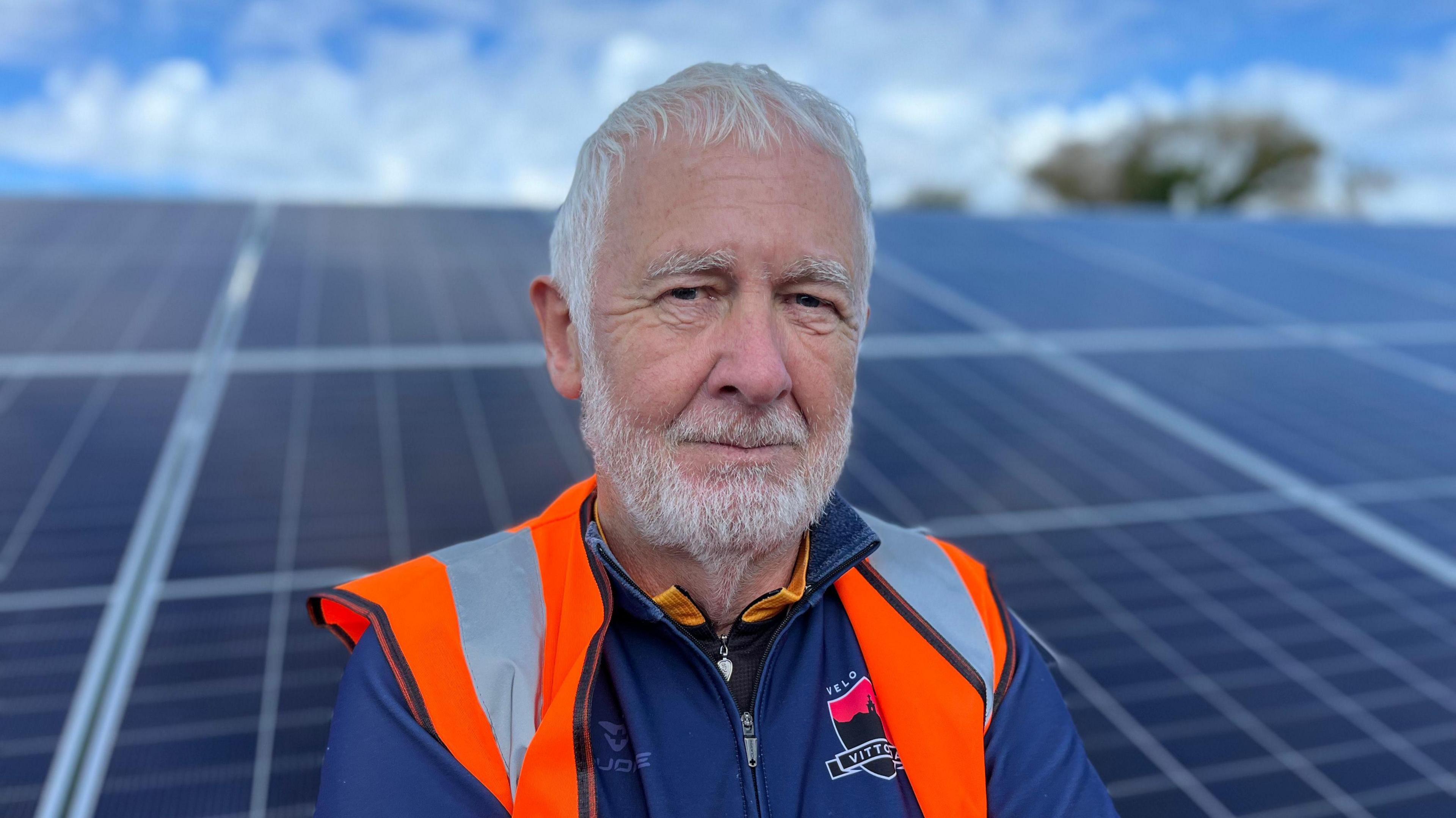 Peter Hargreaves is a white man with white hair looking into the camera. He is wearing a green navy cycling top underneath an orange high-viz vest. The background is an out of focus solar panel and blue sky with clouds.