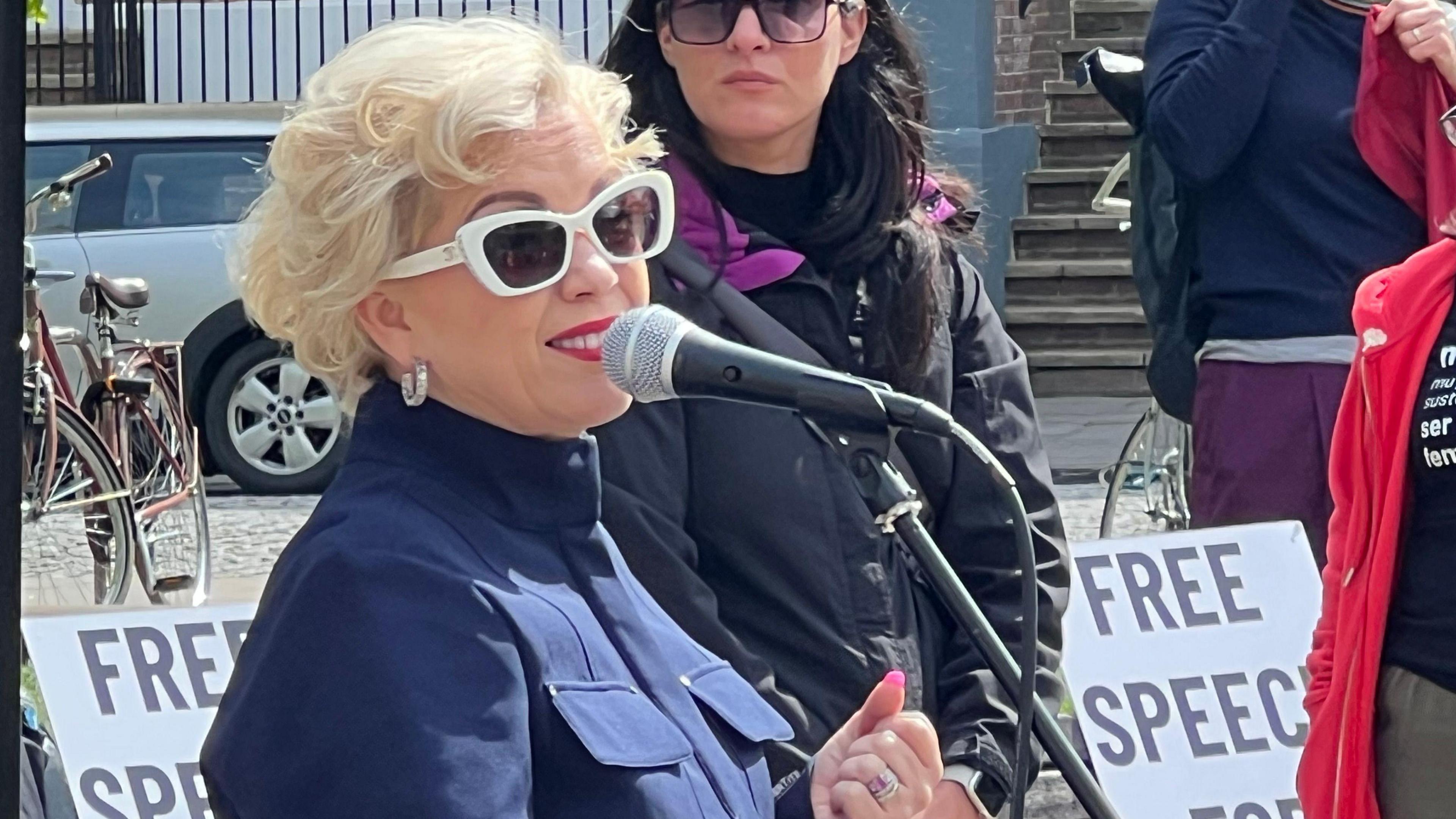 Kellie-Jay Keen wearing white shades and a dark top standing in front of placards and people and talking into a microphone