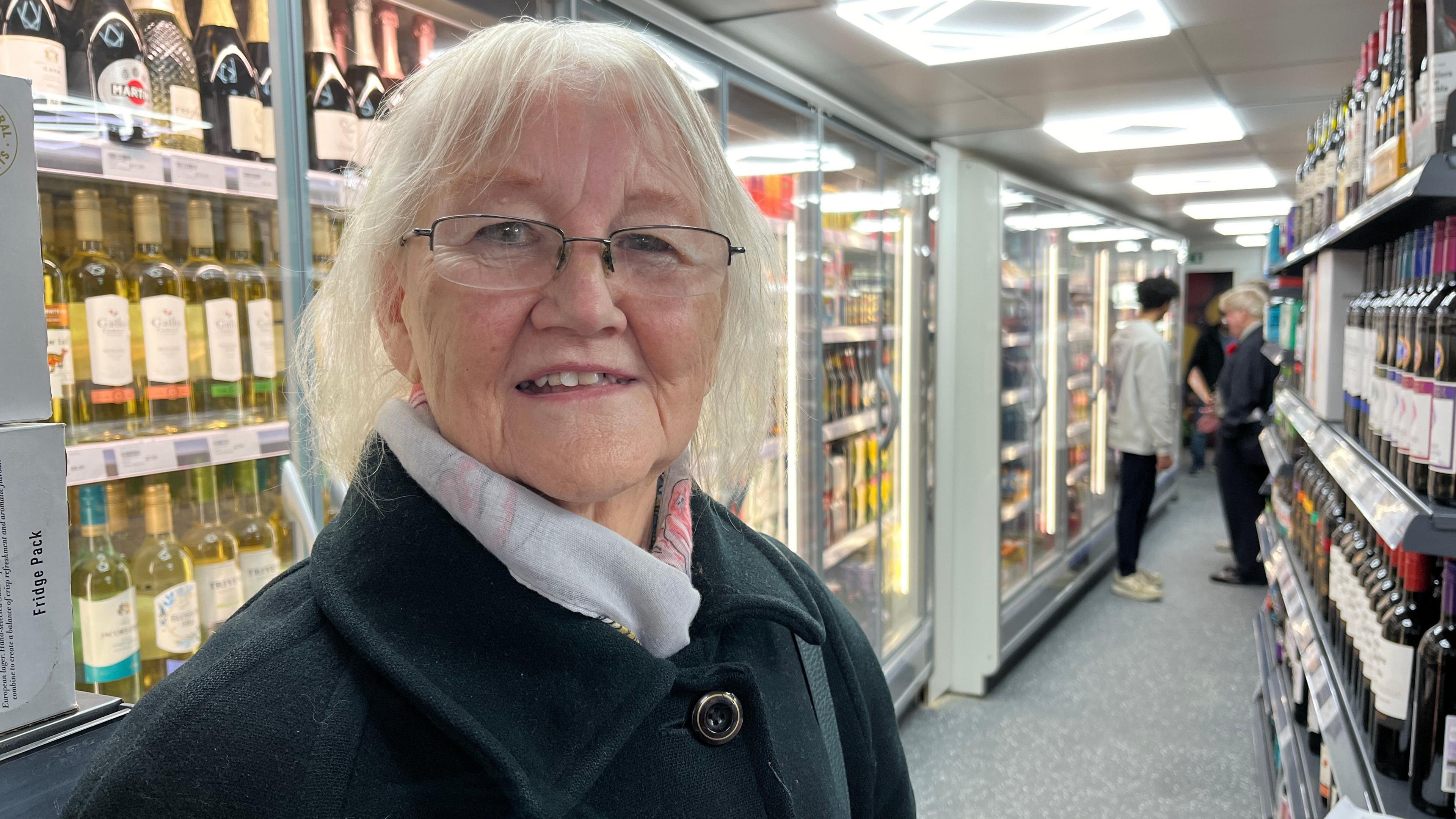 Ms Lacey is standing inside the well lit shop. Shelves on both sides are stocked with bottles of wine and other beverages. On the left, there are multiple rows of white and red wine bottles. She Is wearing a blue coat and has medium length white hair. She is also wearing glasses.