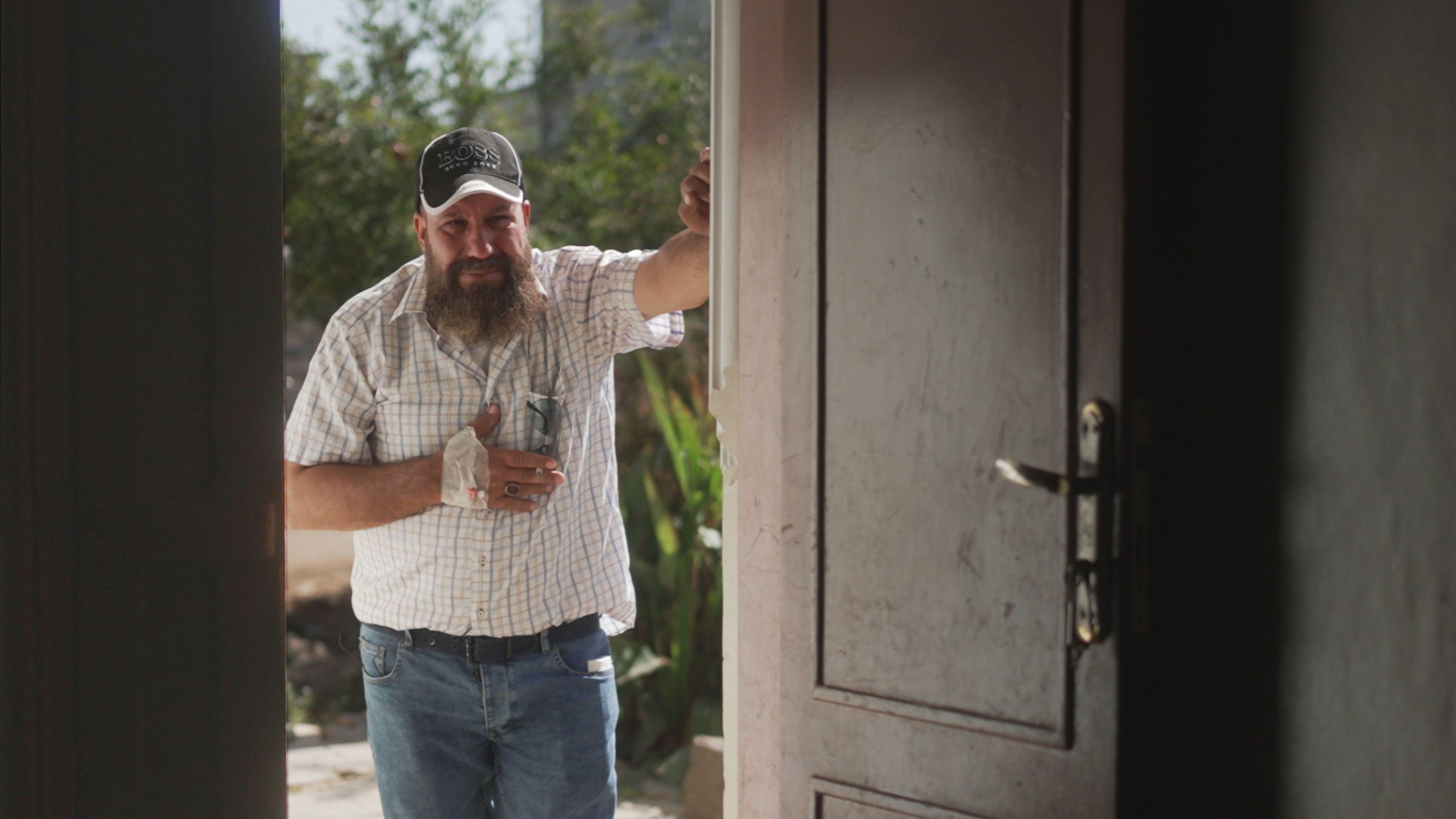 A man is standing in the sunshine in the doorway of a house, looking in towards the camera. He has dark eyes and a long dark beard, which is going grey at the edges, and is wearing jeans and a white shirt. He has a baseball cap on, and is clutching at his chest pocket with his hand, which has a bandage wrapped around it.