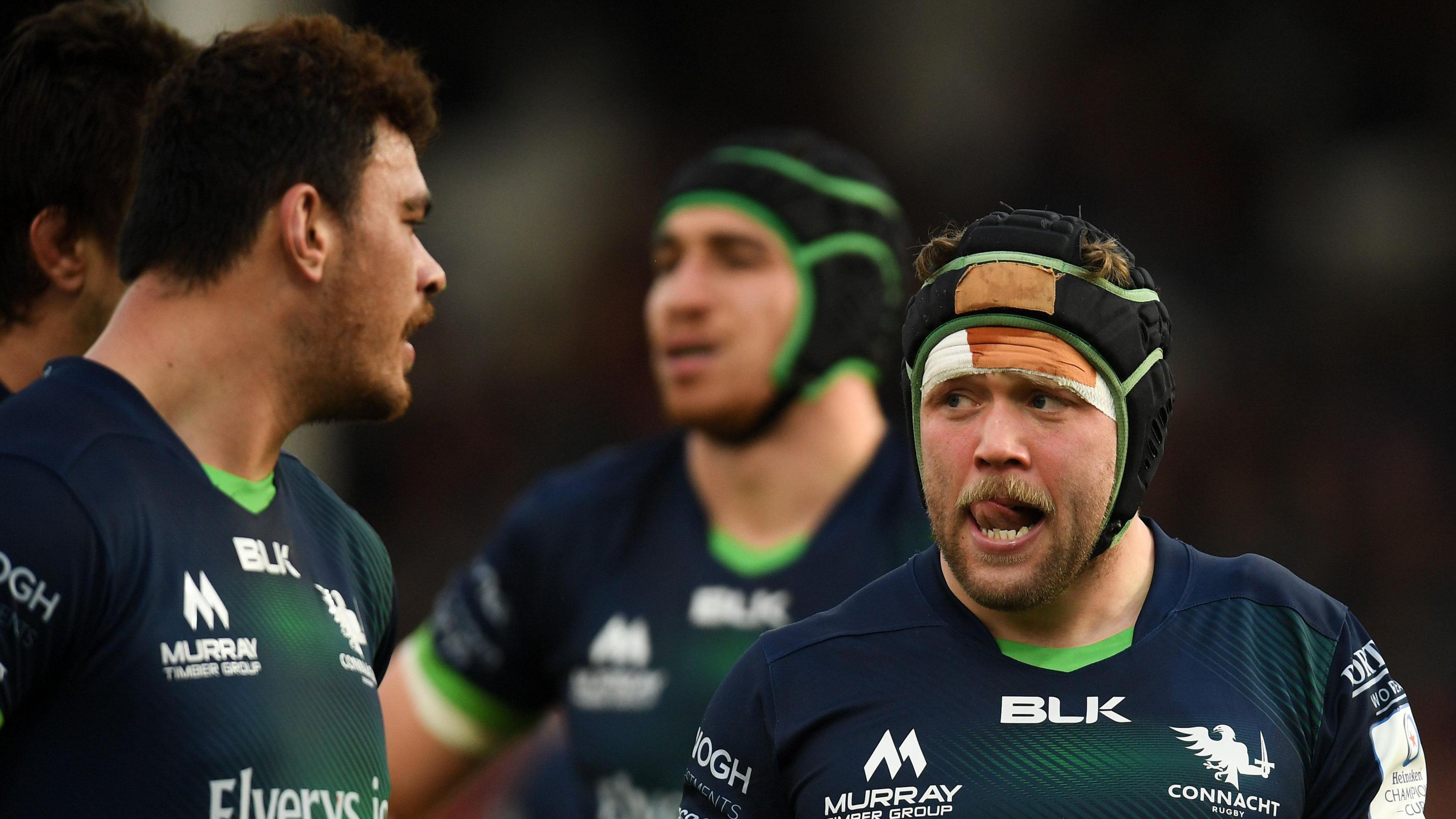 Jonny Murphy right of centre. Wearing a black scrum hat. He is touching his lips with his teeth and looking at his teammate. He is wearing a navy and green coloured Connacht rugby jersey. There are two other players in the background.