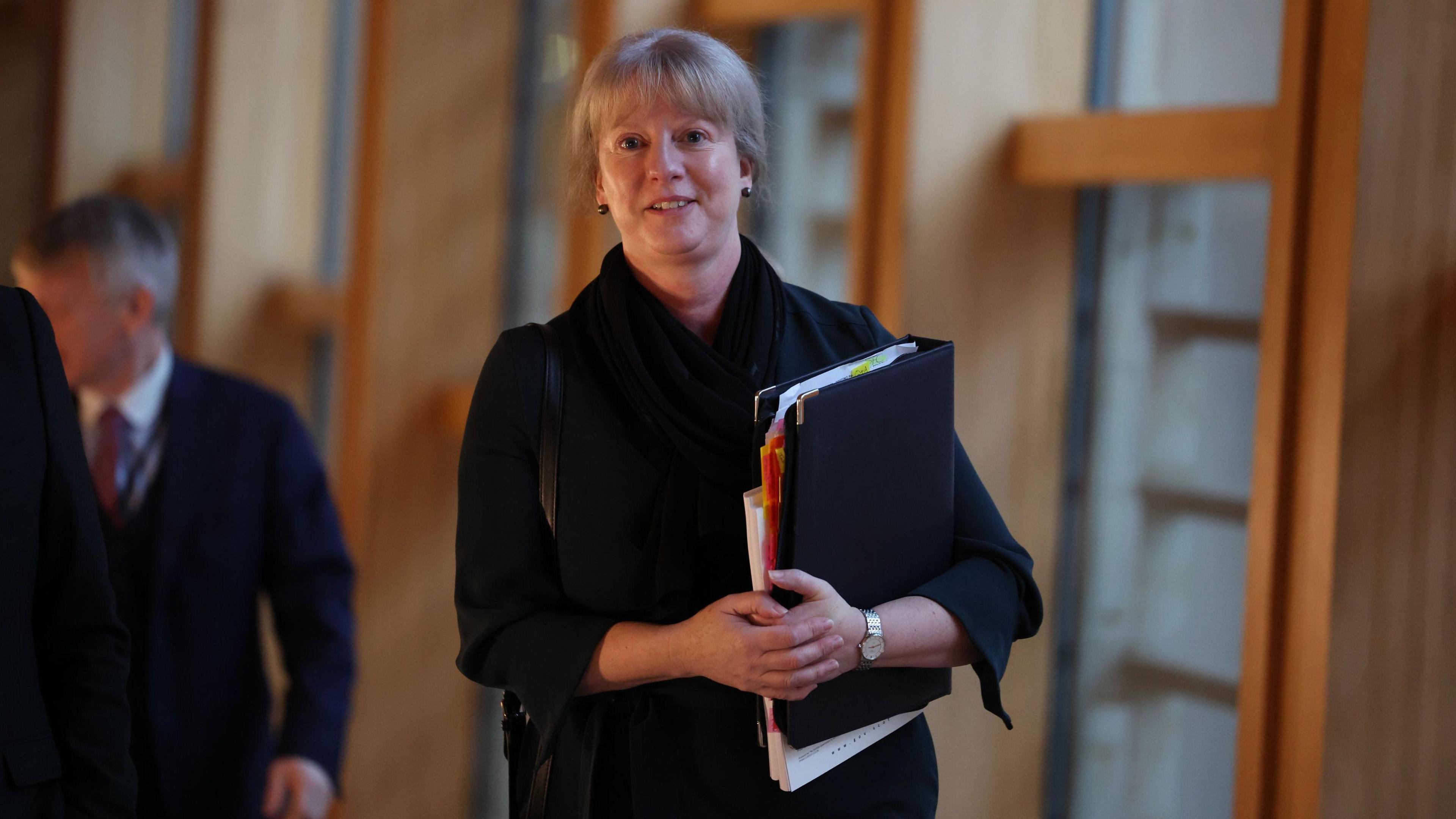 Shona Robison walking in the black and white corridor at Holyrood. She is carrying a binder full of papers and is smiling as she approaches the camera.  She is wearing a dark suit. A man can be seen walking behind her.