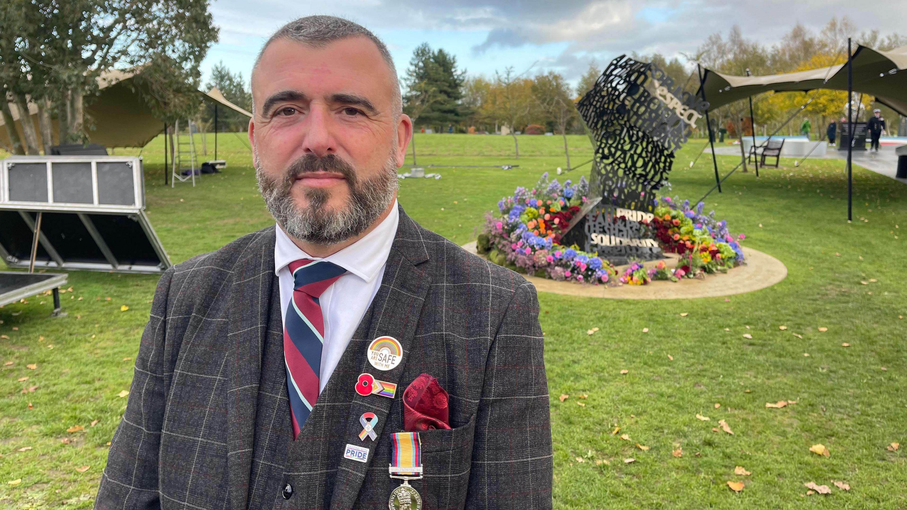 A man with short hair and a beard, wearing a grey checked suit with a red, white and blue tie, pride and remembrance badges and a military medal, stands in front of the LGBT+ armed forces memorial, which resembles an unravelling piece of paper and is surrounded by brightly coloured flowers.