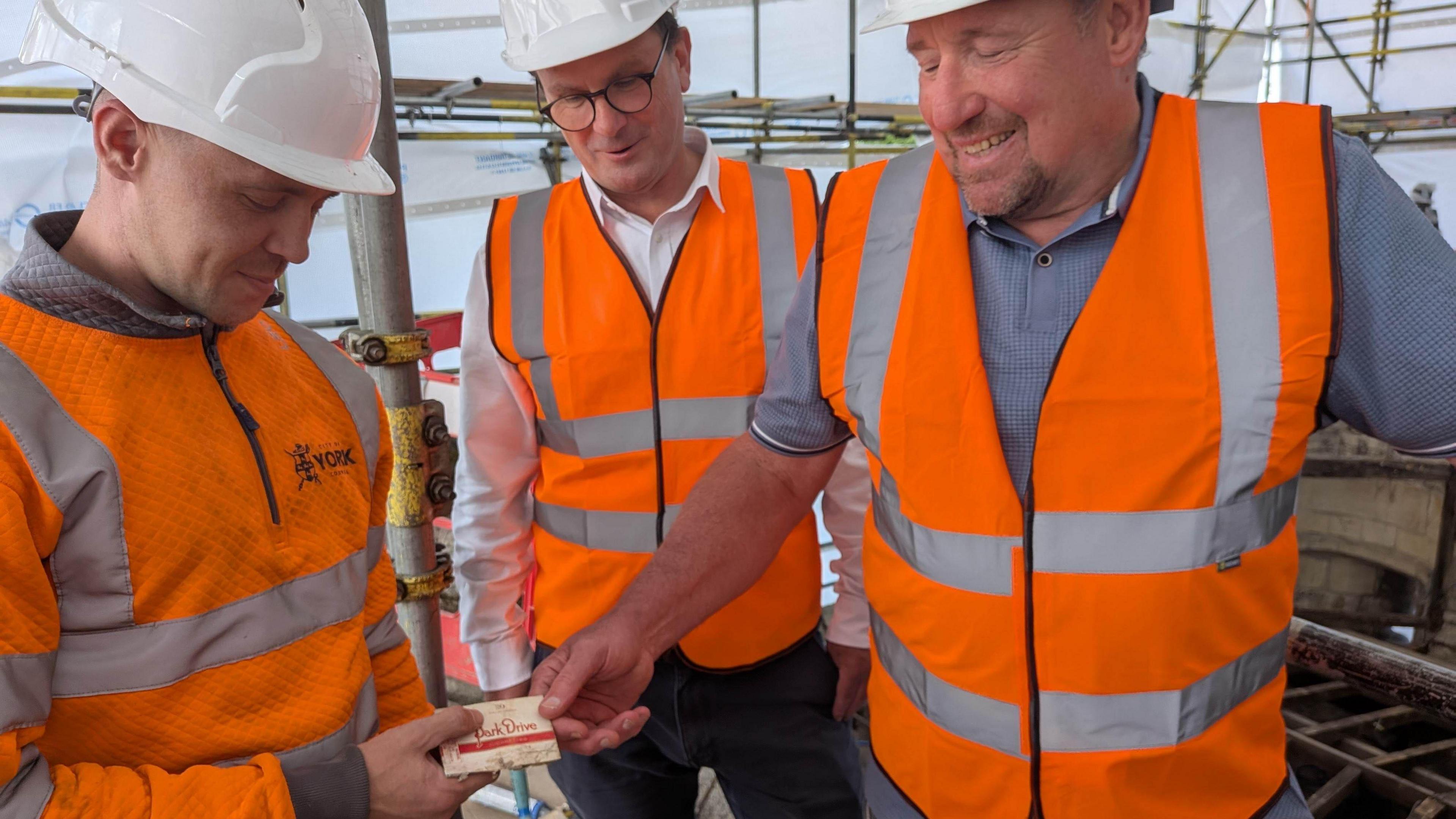 Three men wearing hard hats and orange high-visibility clothing stand on a building site. One hands a cigarette packet to another, who is smiling.