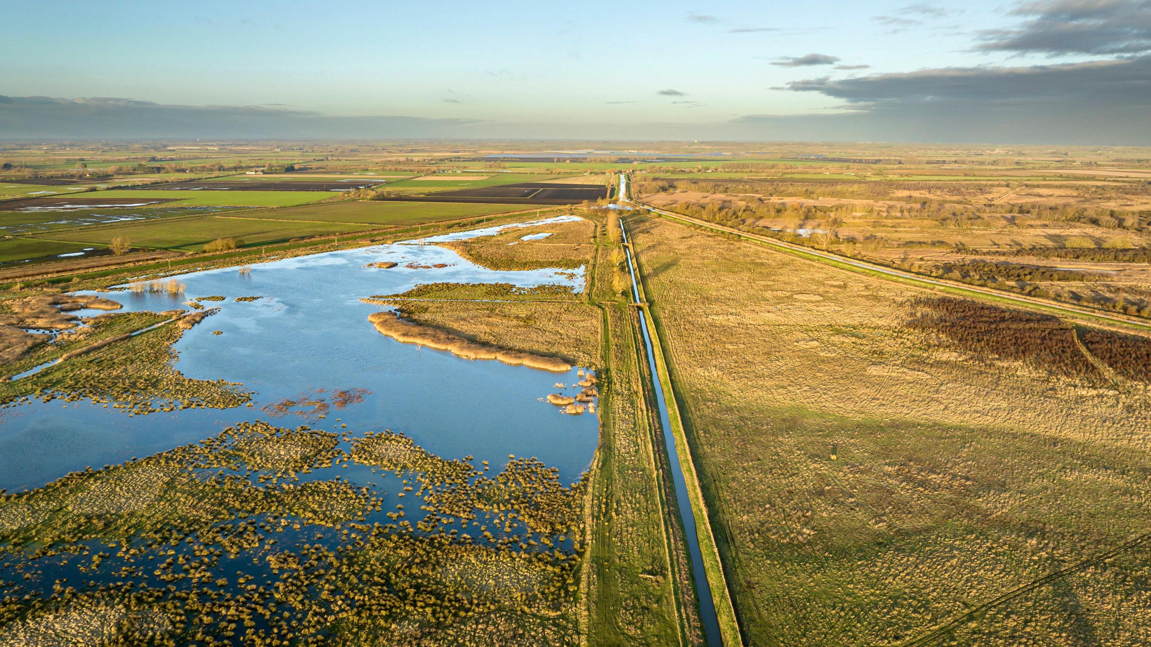 An aerial shot of Wicken Fen nature reserve. It shows a flat green landscape with a large expanse of water on the left. A straight waterfilled drove bisects the view. Above it is a blue sky with hazy clouds. 