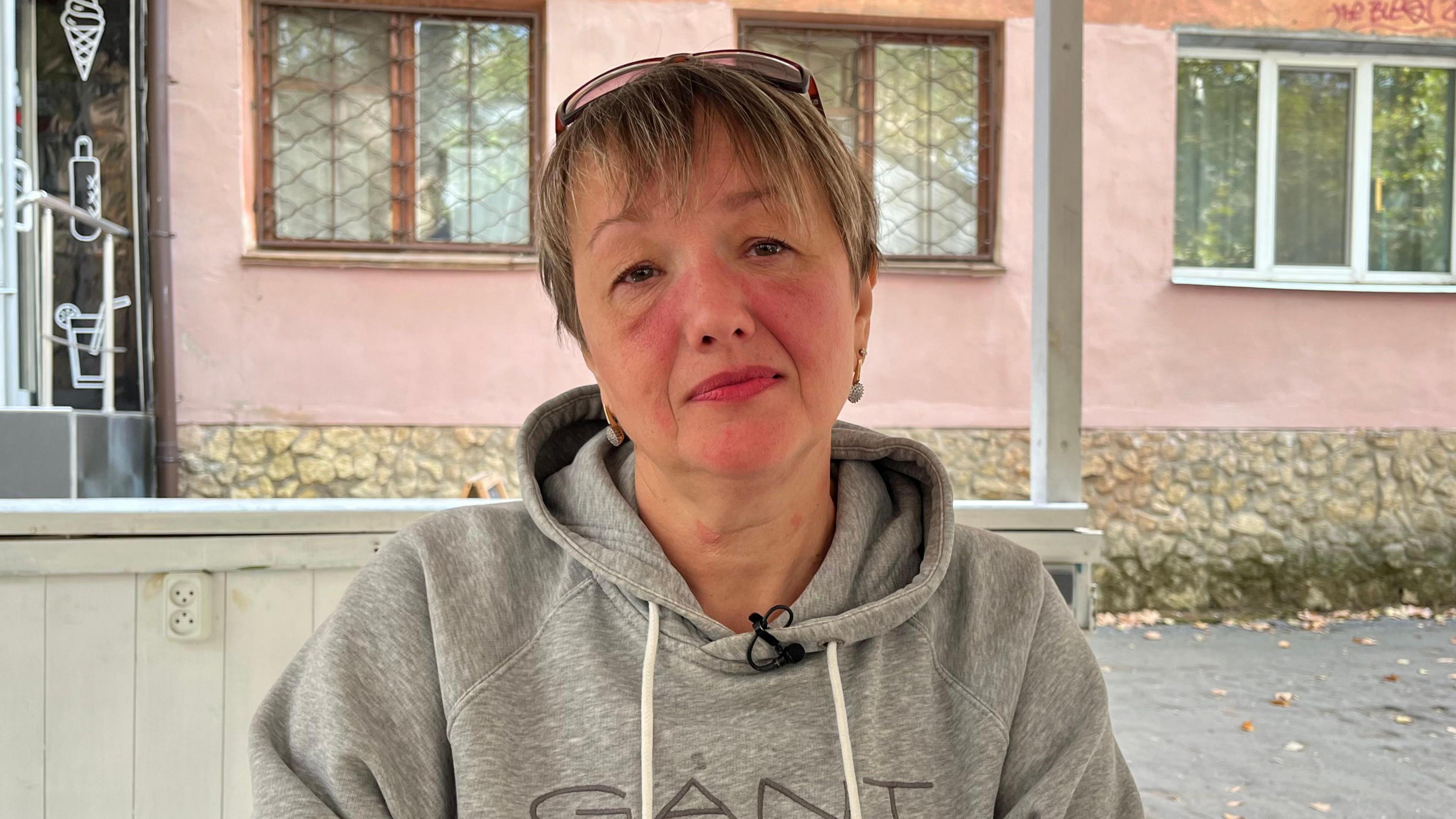 Angela, a woman with short blonde hair and sunglasses on her head, wearing a grey Gant hoodie, faces the camera in an outdoor porch area looking out on a building with pink walls in Kherson