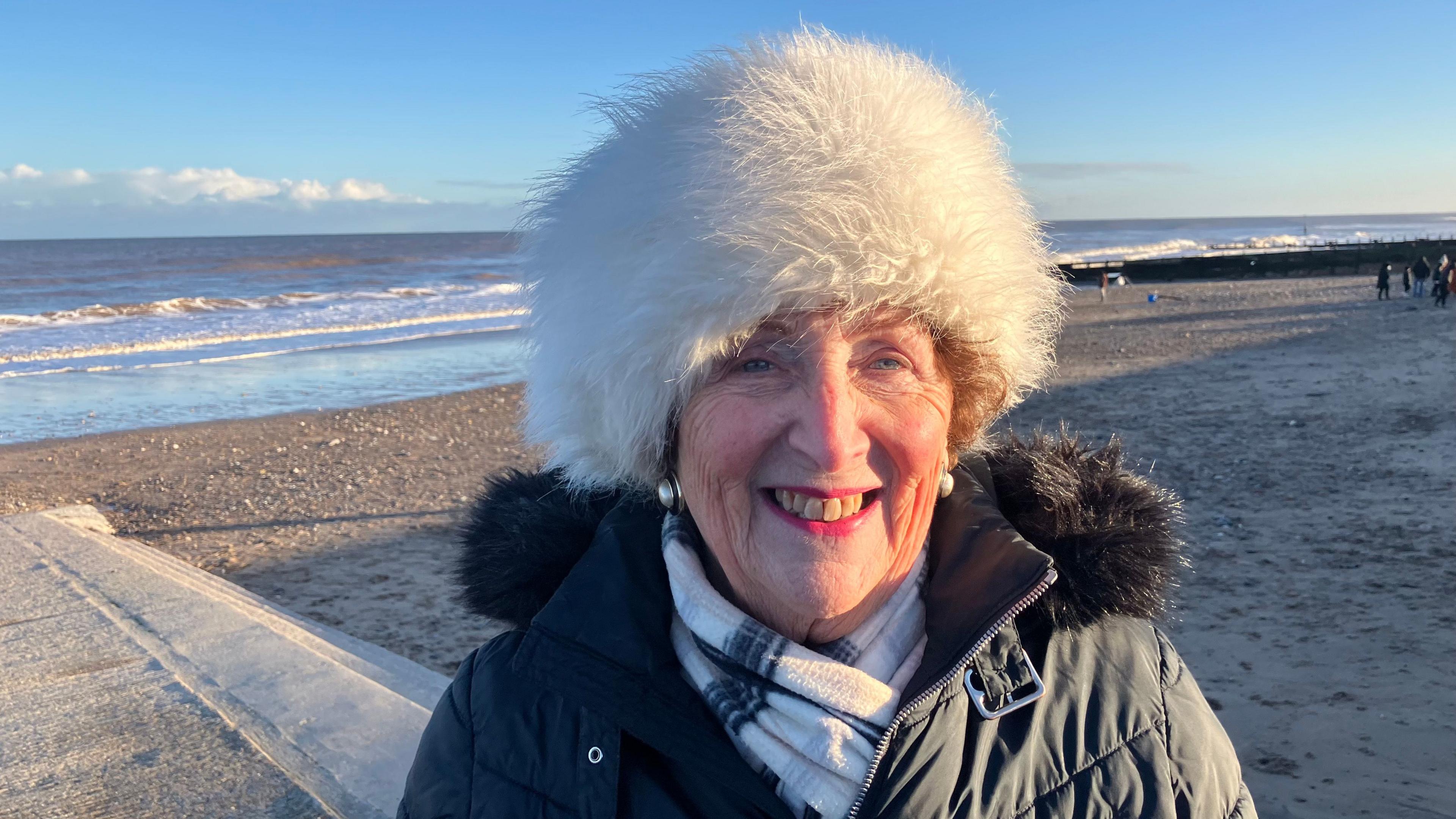 Barbara Jefferson standing on a slipway with the sea in the background. She is wearing a scarf and coat with furry collar, as well as a furry white hat.