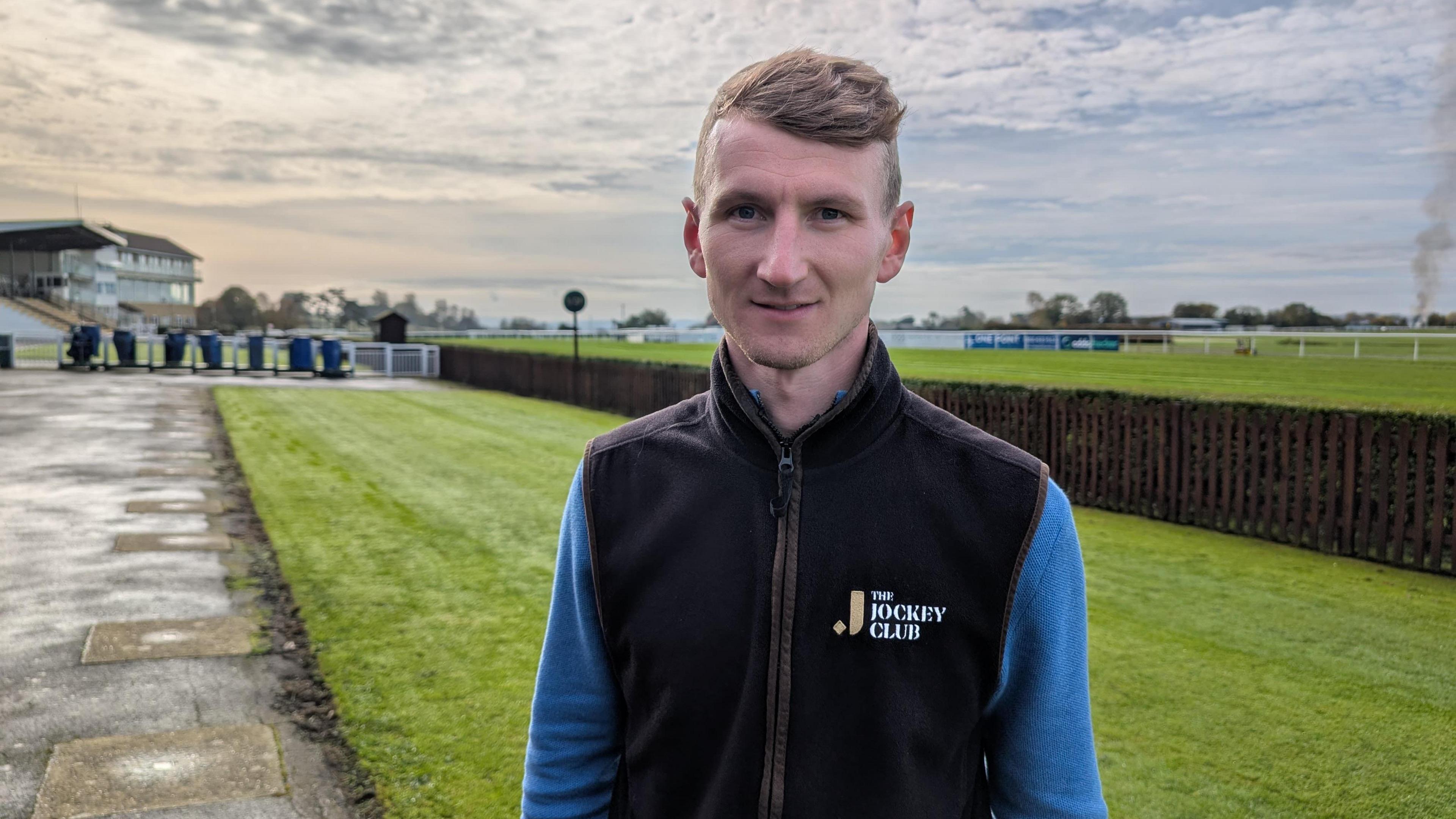 Tom Ryall is standing in the middle of the shot, wearing a blue jumper and black fleece vest on top. He is looking at the camera with a neutral expression. He is standing in front of the racetrack and the stands.