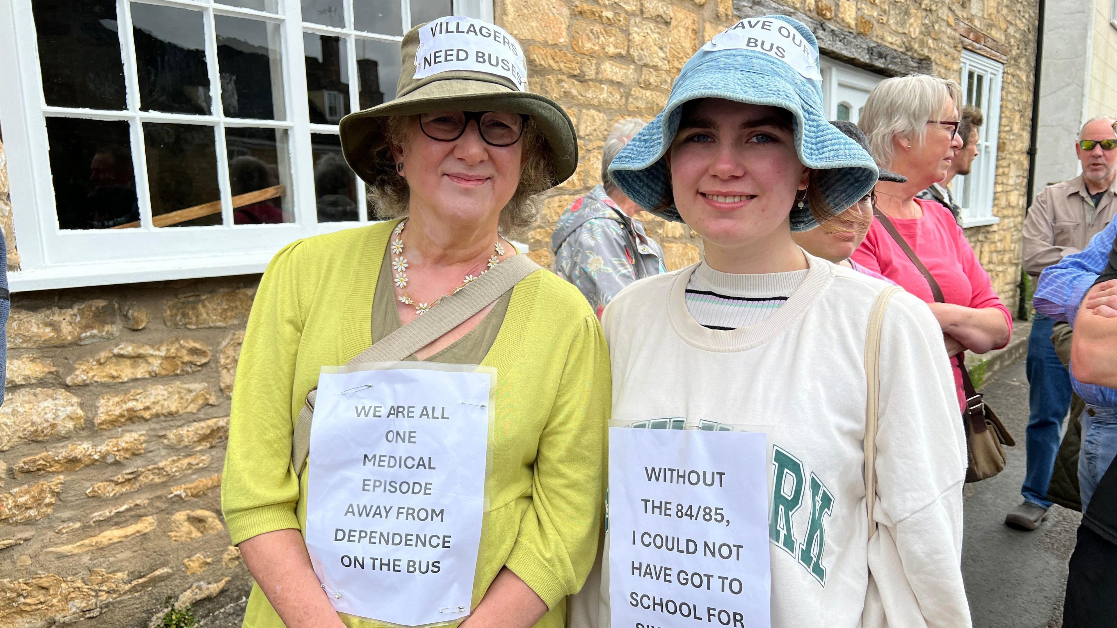 Debbie Young with her daughter, wearing hats and signs expressing their support for the bus route