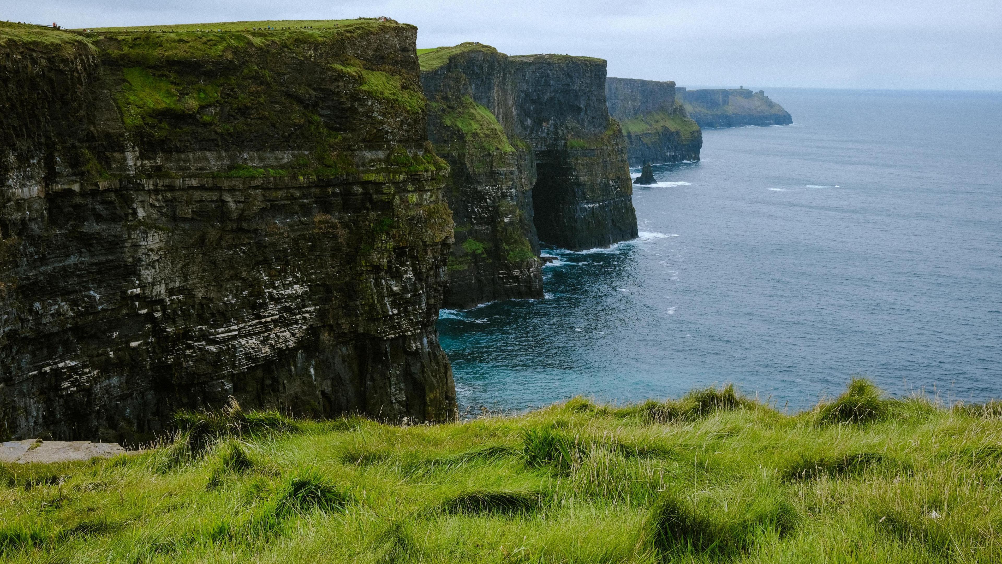 A vista of the Cliffs of Moher. The photo is taken from an edge of grass leading to a drop. The sky is cloudy.