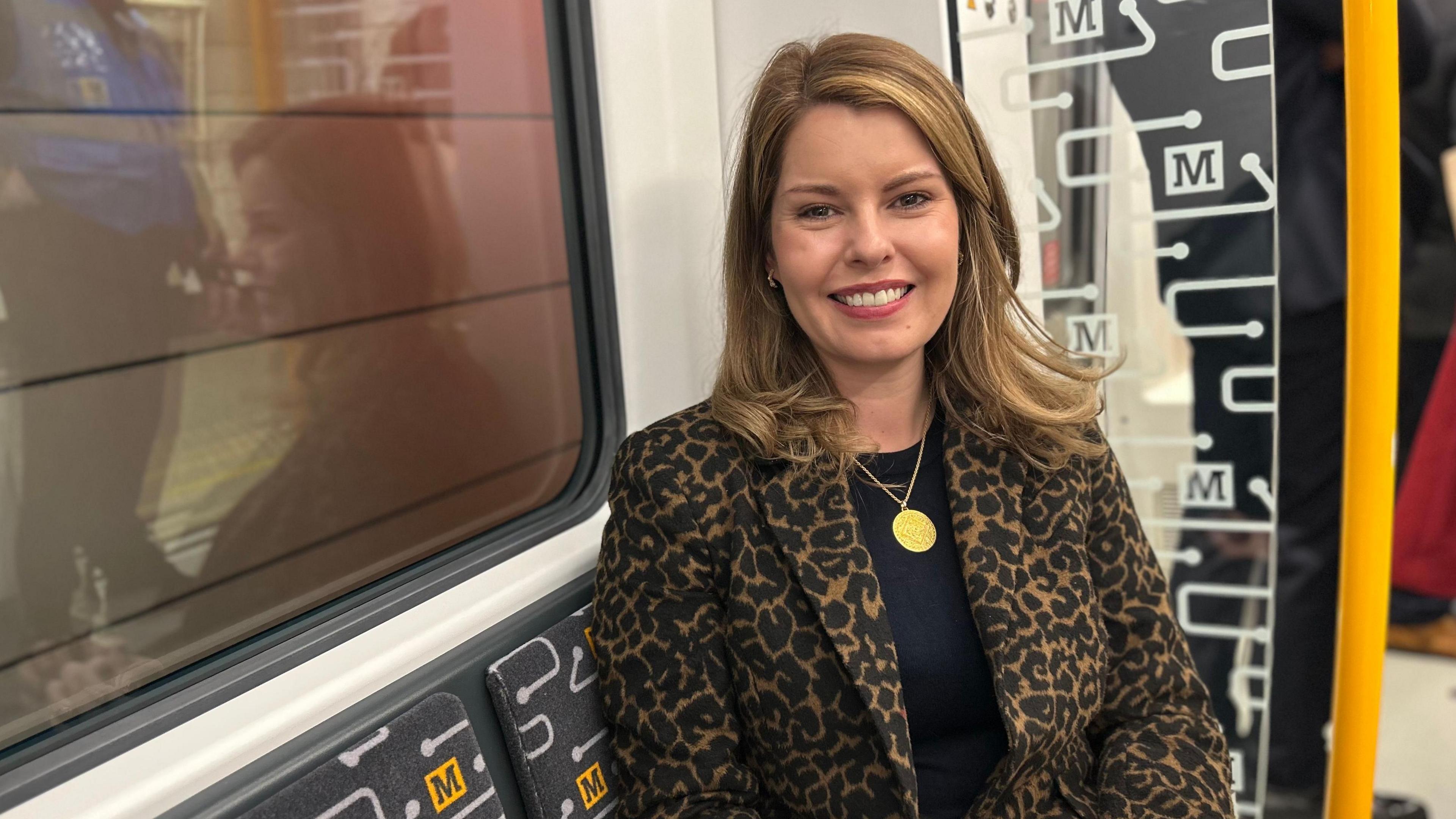 Kim McGuinness is sitting next to a window on a train on the Tyne and Wear Metro. She has blonde hair and is wearing a large gold pendant and is wearing a leopard skin jacket