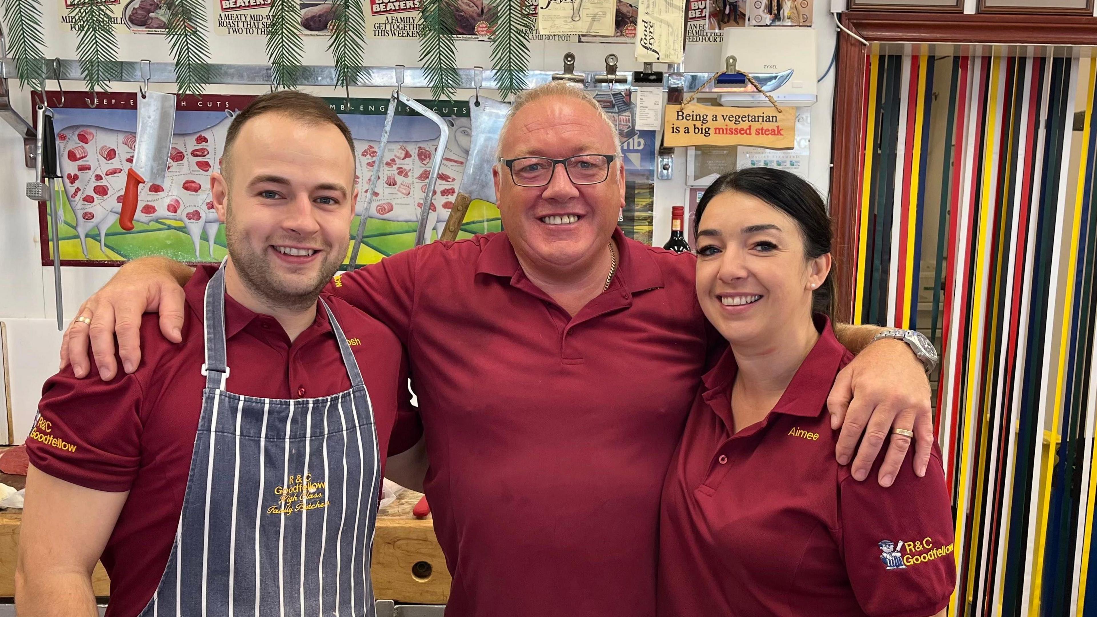 Three people in maroon shirts stand together in a butcher shop, with one wearing a striped apron. Their arms are around each other. Behind them is a selection of meat on a chopping block alongside knives and saws. A door on the right of the image is obscured by colourful ribbons.