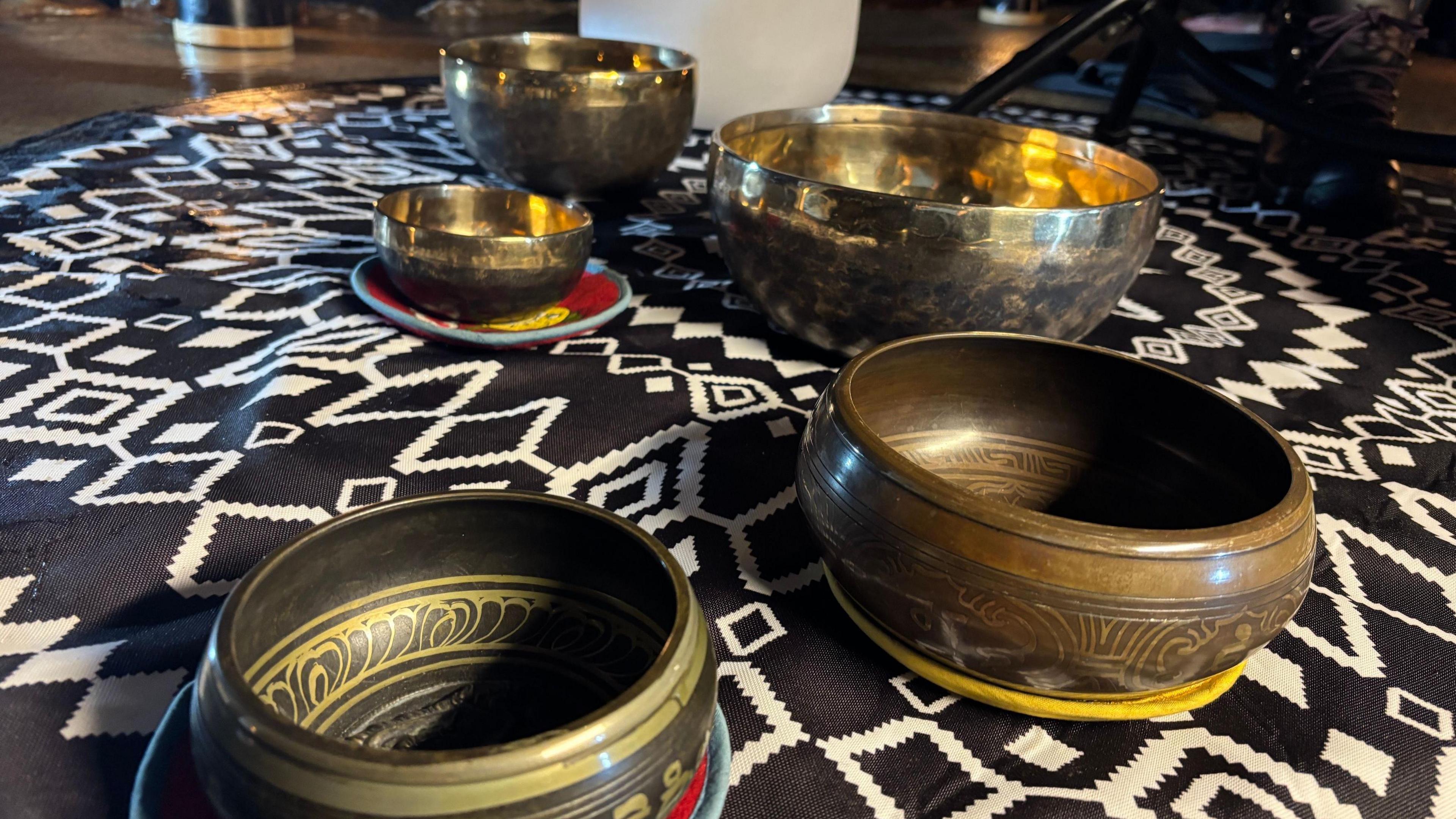 Five bowls sit on a patterned rug in darkness. some of the bowls are shiny metal, some are wooden and some have patterns on.