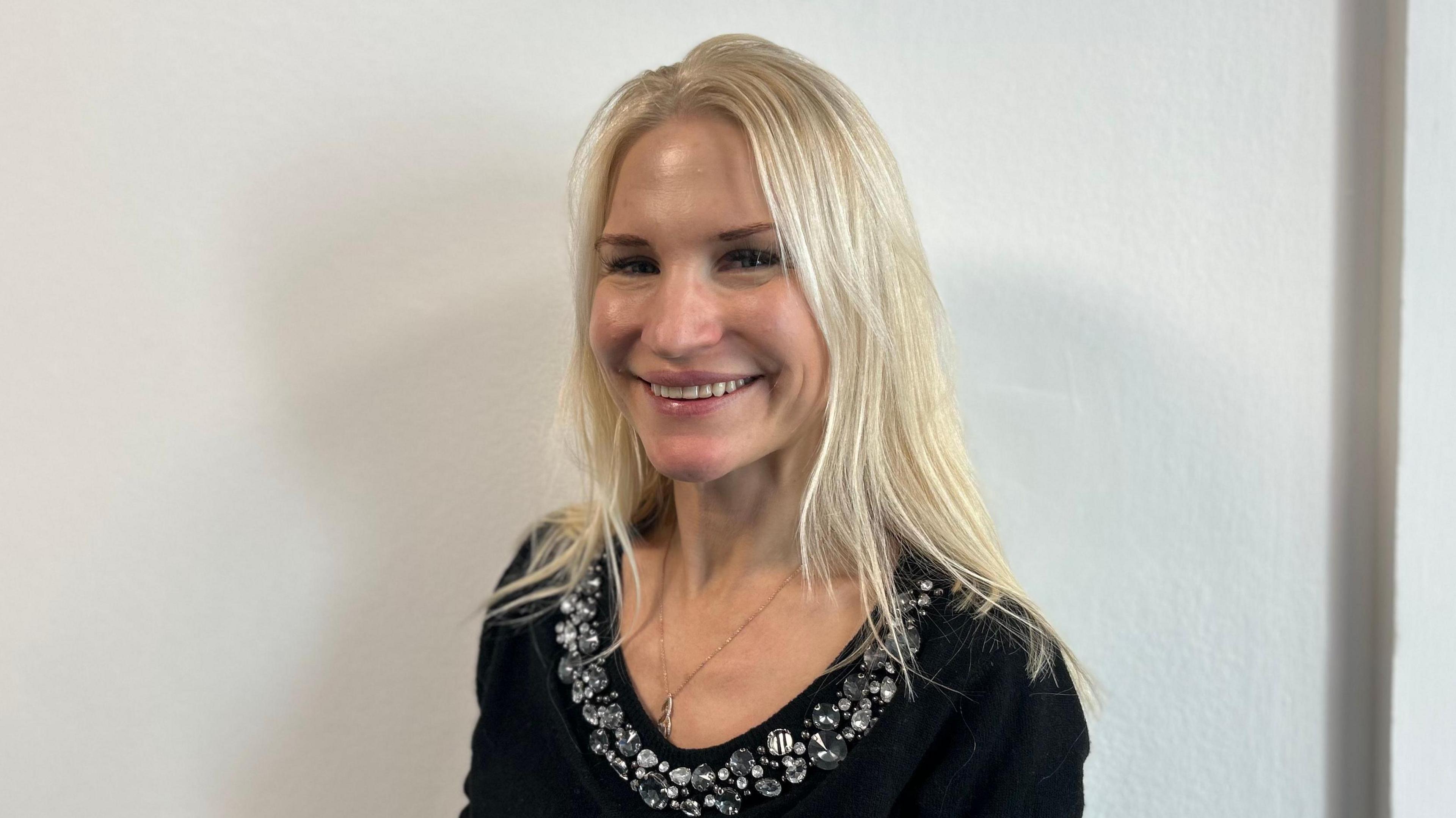 A woman with long blonde hair and wearing a black jumper with a jewelled collar smiling at the camera against a white background.