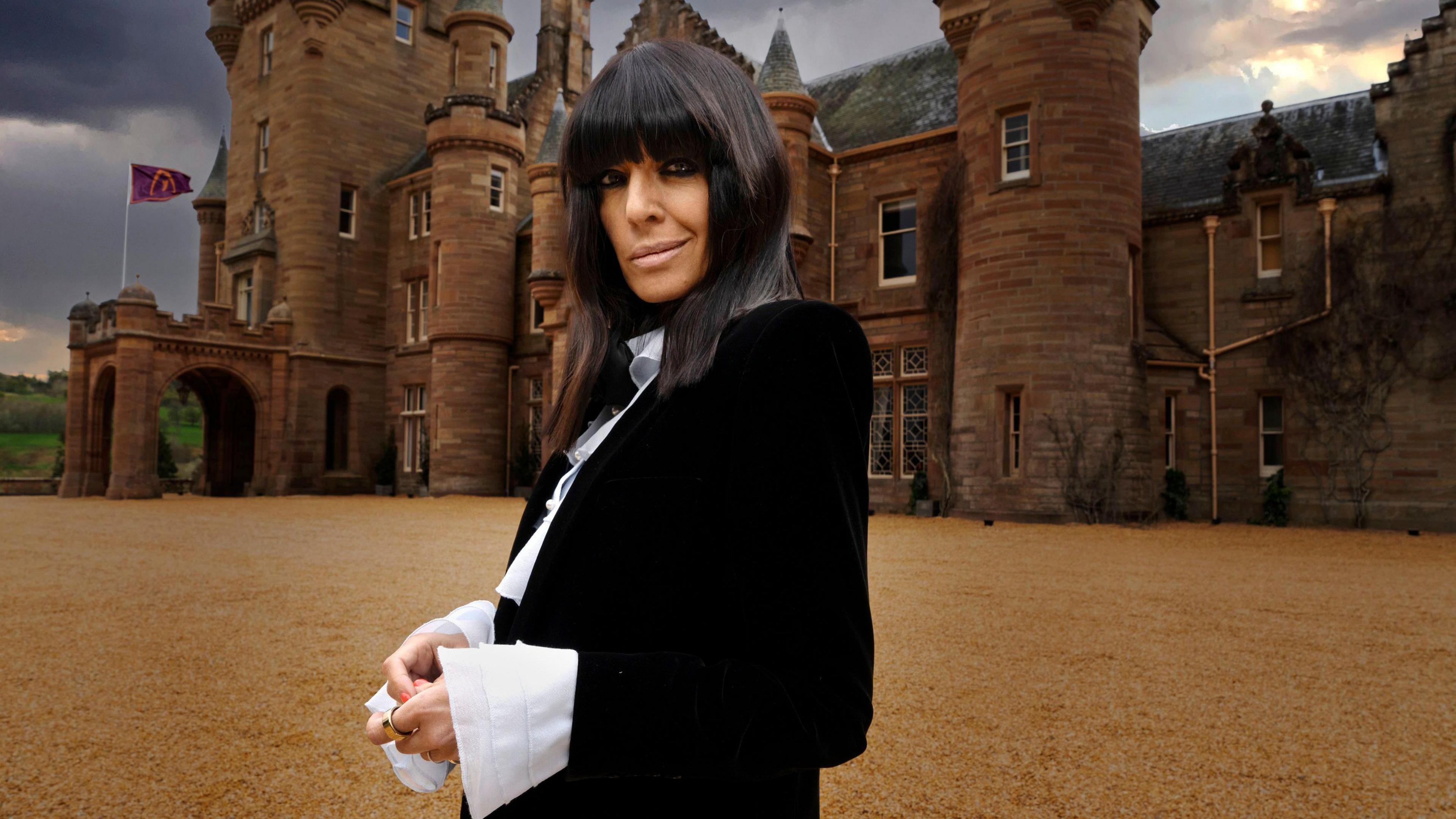 Claudia Winkleman wearing black and standing in front of a castle. 