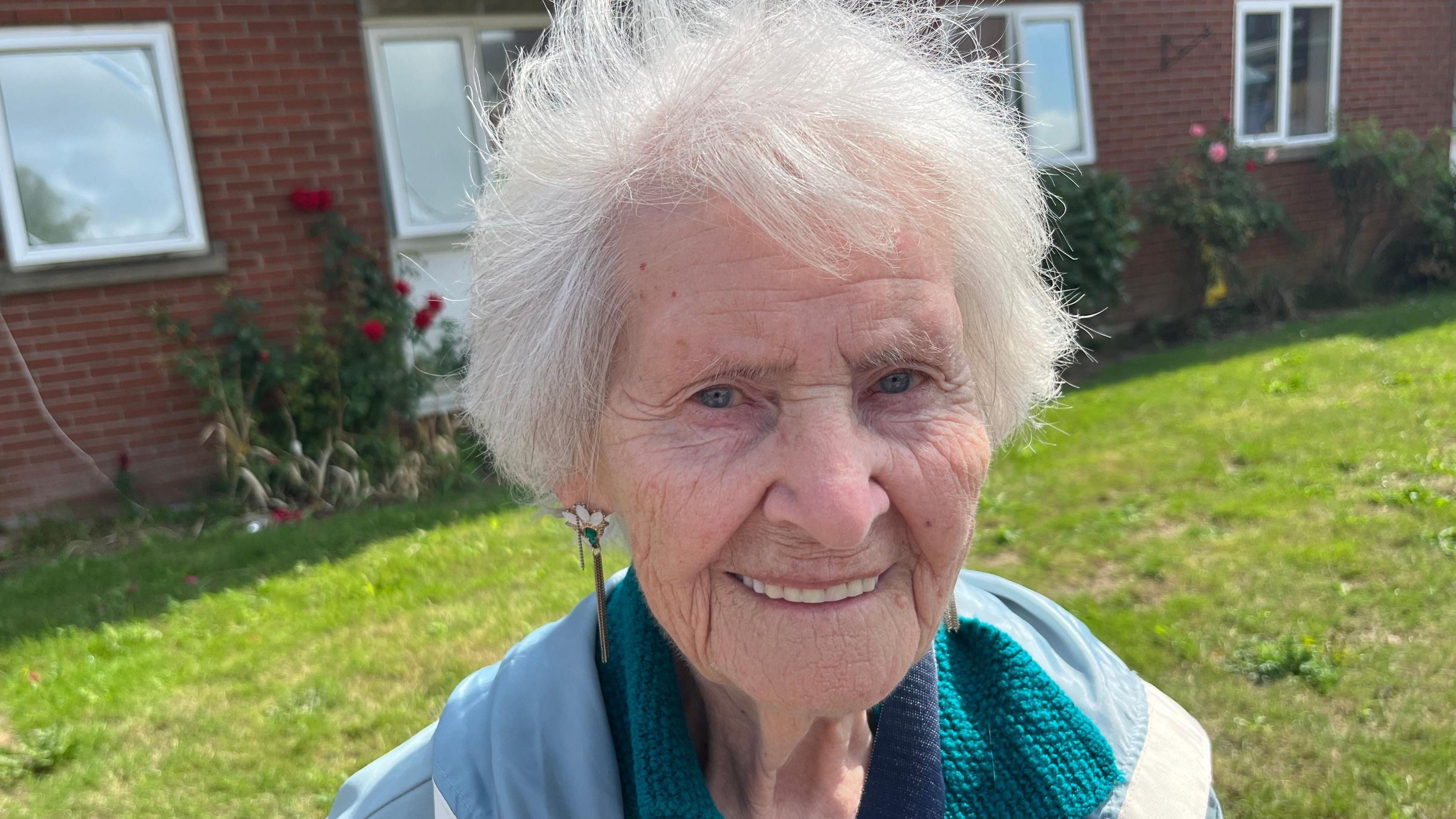 Joyce Chaffe is smiling at the camera while standing outside some of the homes. She has short white hair, blue eyes and dangly earrings. She is wearing a blue coat and the sun is shining on the grass behind her.